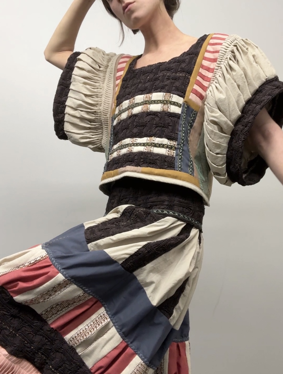 A figure wearing a sculptural garment composed of hand-pieced and pleated cotton in black, cream, and striped panels of red, navy, and gold. The top features puffed sleeves with elaborate ruching and embroidered black textile bands accented with cream ribbons, while the lower section displays voluminous pleated skirt panels in contrasting colors. The intricate construction creates dramatic three-dimensional form and texture against a neutral background.