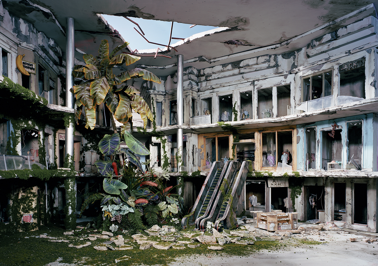 A deteriorating indoor shopping mall overtaken by lush vegetation, with escalators descending into debris-strewn floors. Crumbling concrete balconies, broken windows, and peeling paint frame towering tropical plants and vines reclaiming the abandoned commercial space beneath a partially collapsed ceiling.
