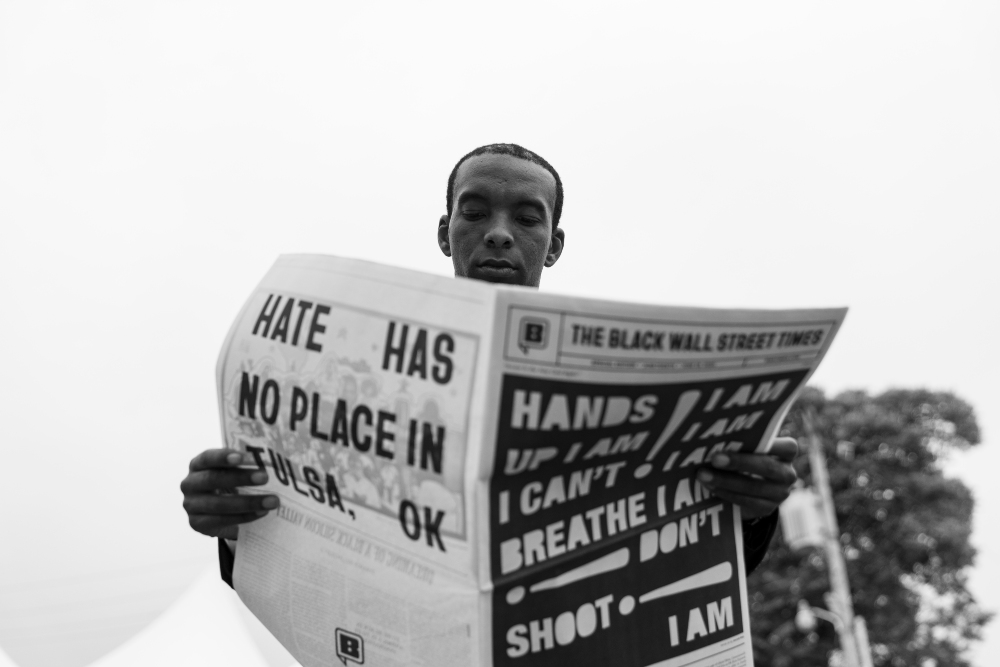 Black and white photograph of a young man holding up a newspaper with protest headlines including "Hate Has No Place in Tulsa, OK" and "I Can't Breathe." The composition is shot from a low angle against a light sky, with trees visible in the background.