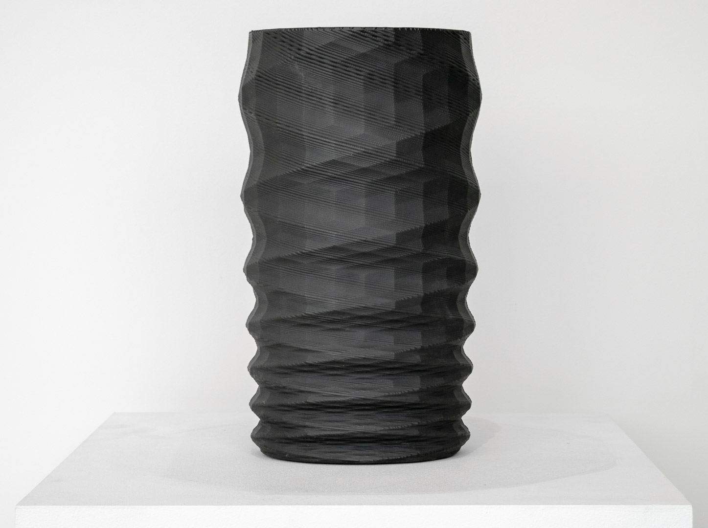 A tall cylindrical black porcelain vessel with undulating, wave-like horizontal ridges along its entire surface, displayed on a white rectangular platform against a neutral background.