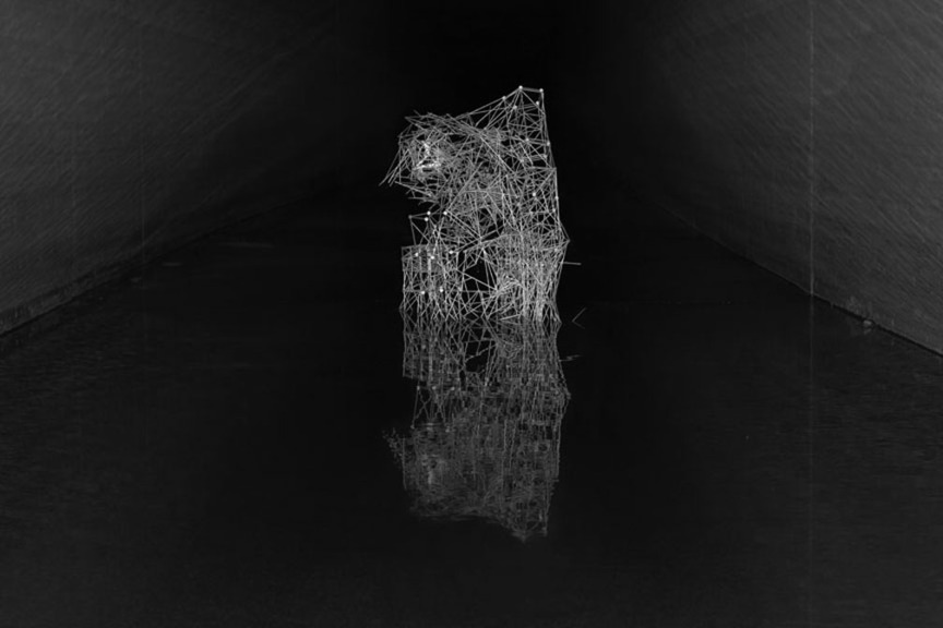 White linear wireframe structure resembling an abstract organic form or architectural element, suspended against a dark background. The densely layered lines create a three-dimensional sculptural silhouette with openwork sections and solid areas, photographed in high contrast black and white.