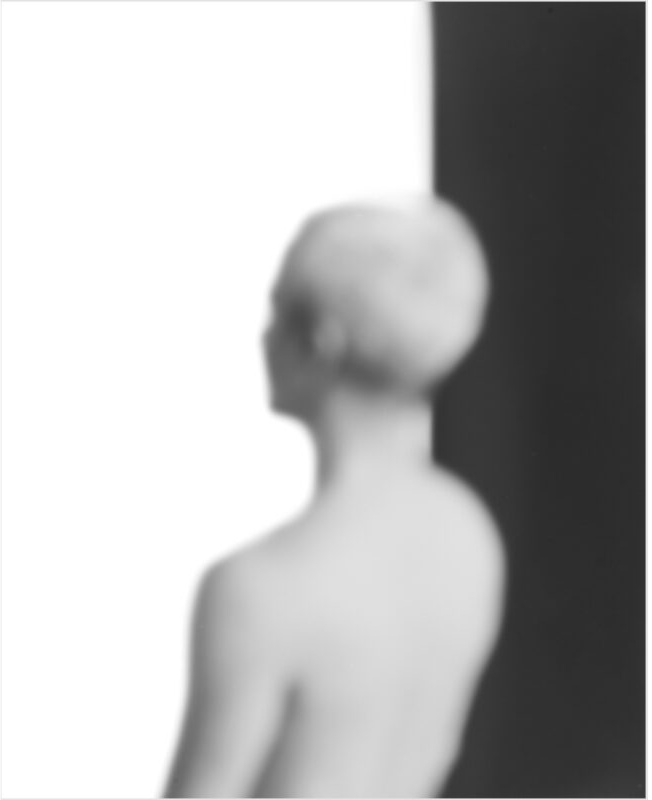 A soft-focus photograph of a white plaster or stone bust of a human head and shoulders shown in profile against a stark division of light and dark backgrounds, creating a minimalist composition that emphasizes the sculpture's gentle forms and tonal gradations.
