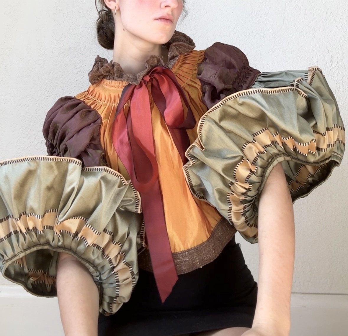 A figure wearing an elaborate sculptural garment with voluminous pleated sleeves in cream and tan tones, paired with a burnt orange bodice and deep red ribbon bow at the neckline. Dark brown puffed shoulders and ruffled brown collar detail complete the richly textured, dimensional composition against a white background.