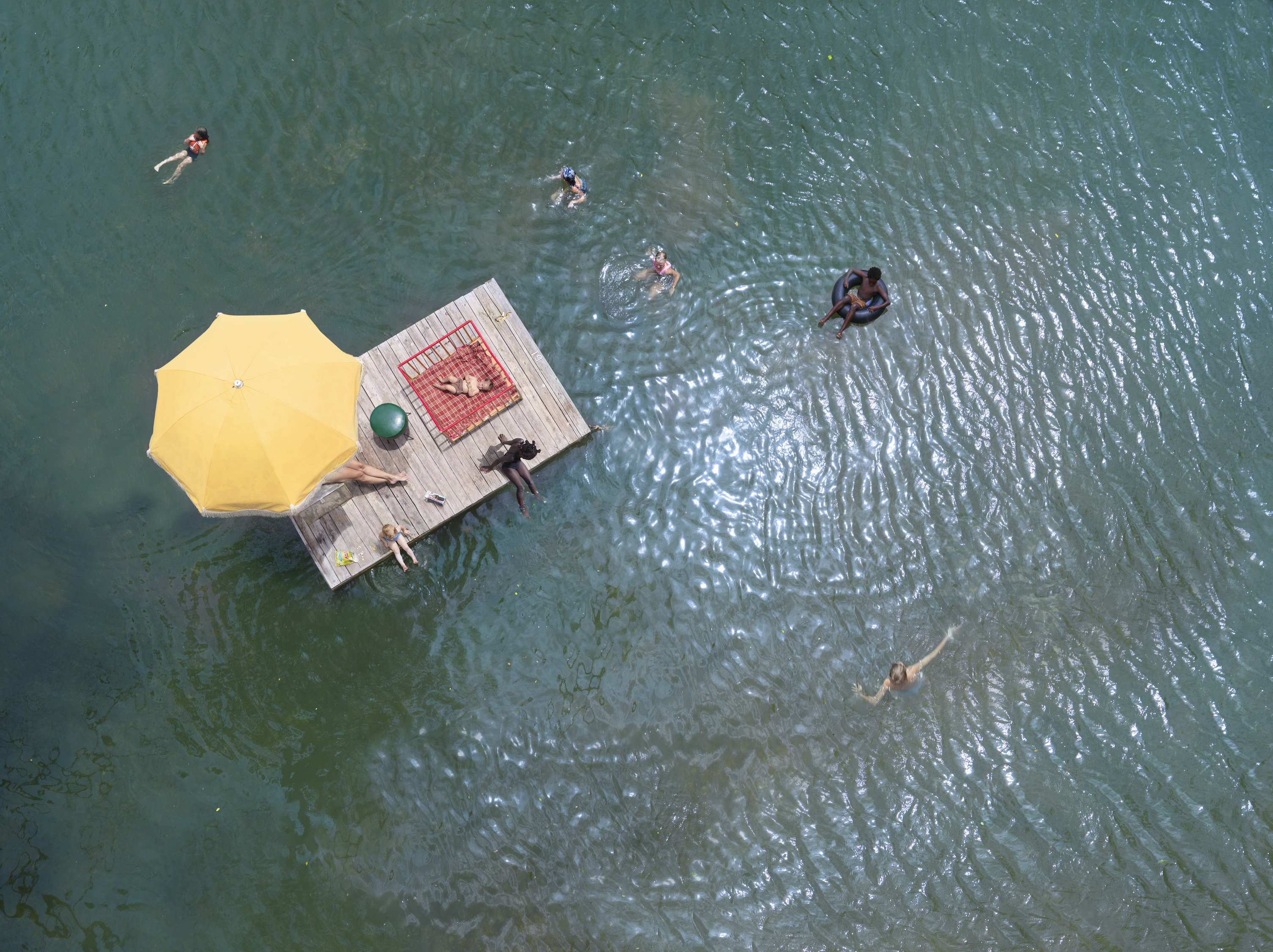 Aerial view of a wooden dock floating on turquoise water, featuring a bright yellow umbrella and a red checkered blanket. Several swimmers in casual summer attire are scattered in the surrounding water, some on the dock and others swimming nearby, creating a leisurely lakeside scene.