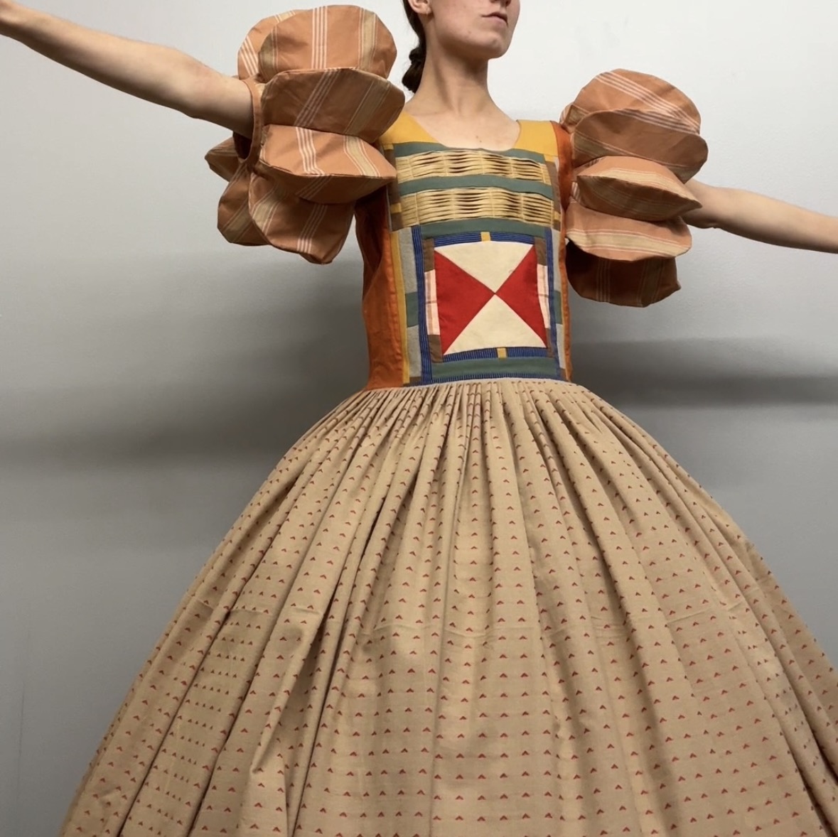 A figure in an elaborate costume stands with arms extended, wearing a voluminous pleated skirt in cream with tiny repeated patterns and a structured bodice featuring geometric patchwork in red, cream, navy, and gold tones. Large sculptural shoulder pieces in striped brown and tan fabric extend outward, creating a dramatic silhouette against a neutral background.