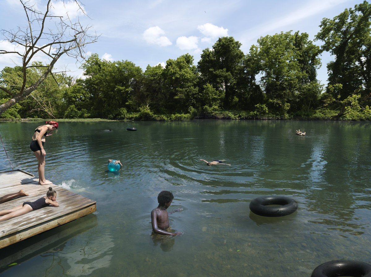 Multiple figures swim and wade in a calm, green-tinted river lined with dense trees under a bright sky. Wooden docks with lounging figures occupy the left foreground, while black inner tubes float in the water among the swimmers.