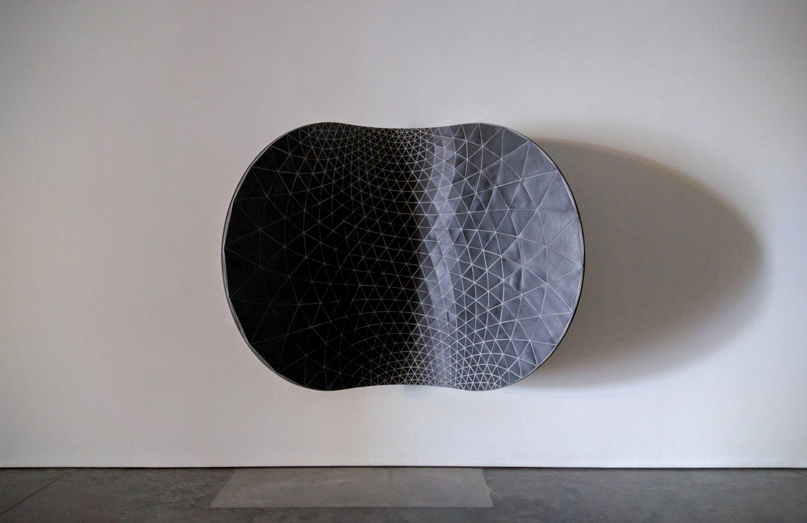 A curved, pod-like sculptural form mounted on a white gallery wall, featuring a densely patterned surface of small triangular tiles that gradient from deep black on the left to light gray on the right, casting a distinct shadow on the wall below.