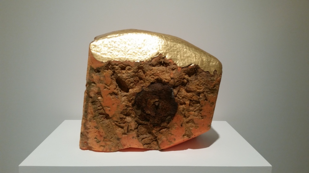 A sculptural form composed of reddish-brown textured material with an irregular, eroded surface sits on a white platform. The top half is covered in burnished gold leaf, creating a stark contrast with the rough, debris-like lower portion.