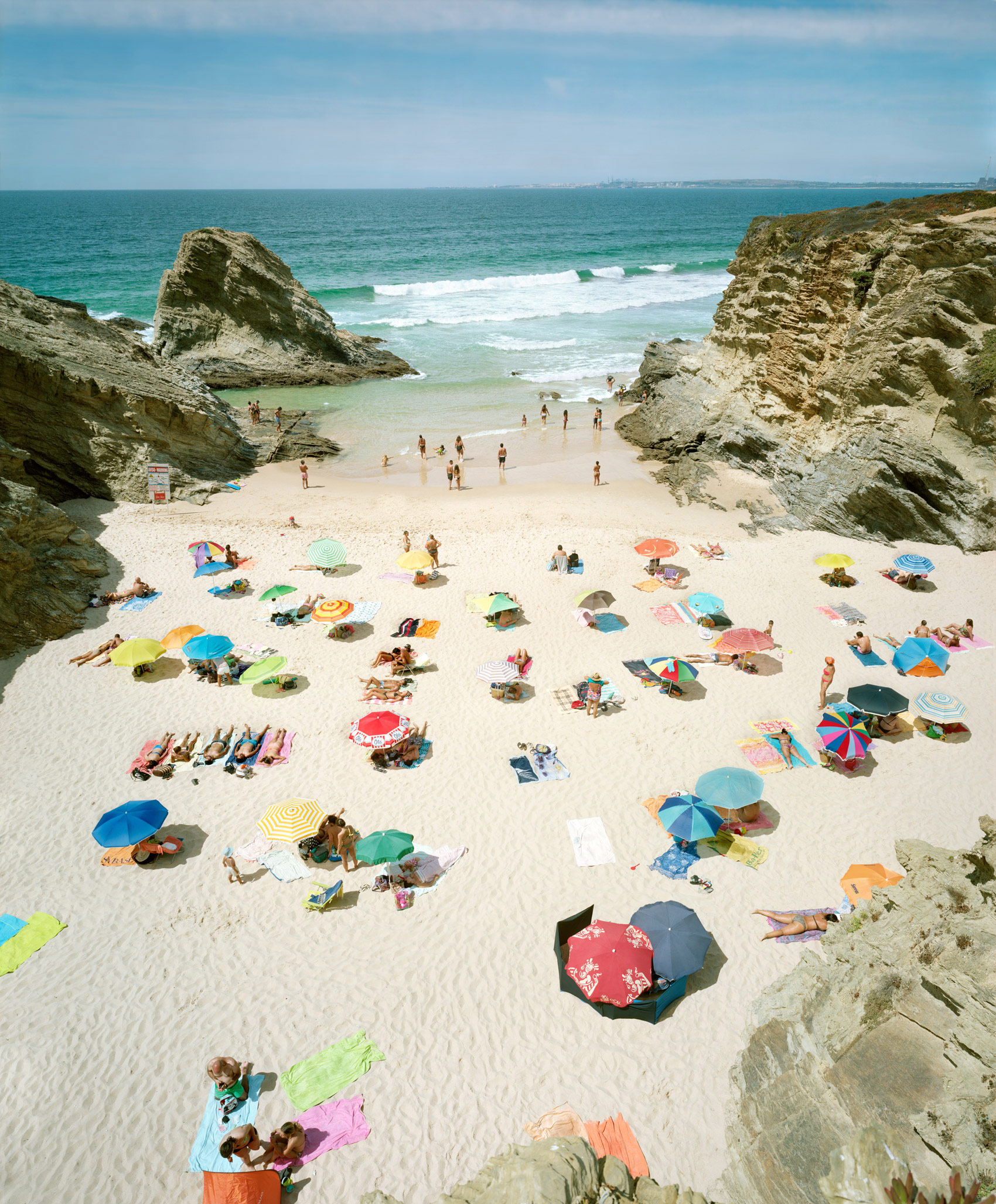 Aerial view of a crowded beach nestled between towering limestone cliffs, with dozens of colorful umbrellas scattered across pale sand and beachgoers swimming in turquoise waters dotted with white waves.