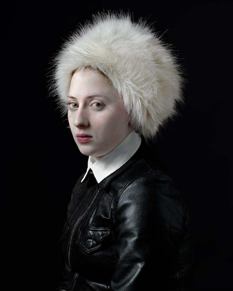 Portrait of a person with pale complexion wearing a voluminous white fur hat and black leather jacket with white collar against a dark background, shown in three-quarter view facing left.