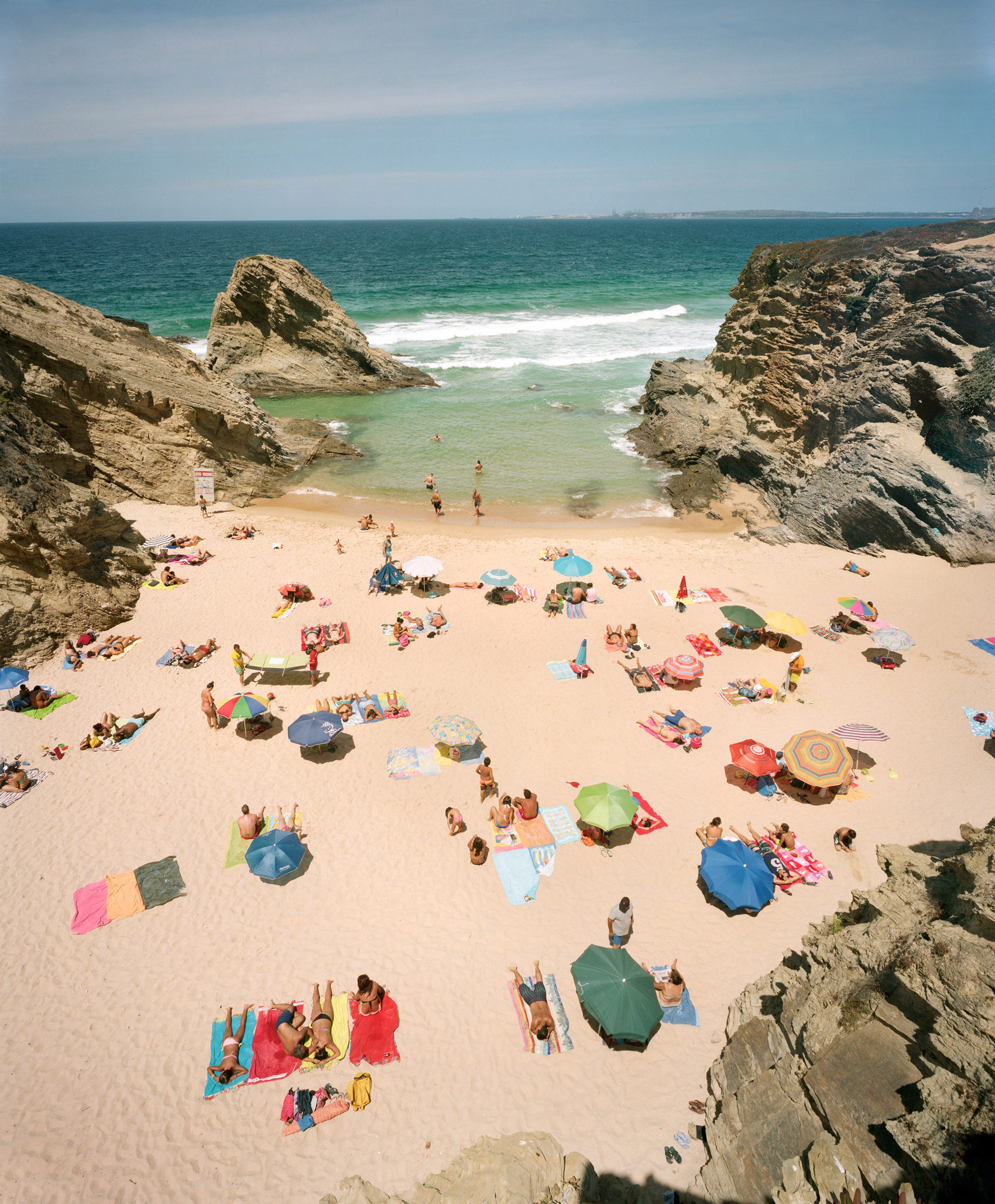 Aerial view of a crowded beach nestled between two large rocky outcrops, with colorful umbrellas and beach towels scattered across the sand. Turquoise water and white-capped waves frame the cove, with beachgoers swimming and lounging in vivid clusters of pink, blue, green, and yellow.