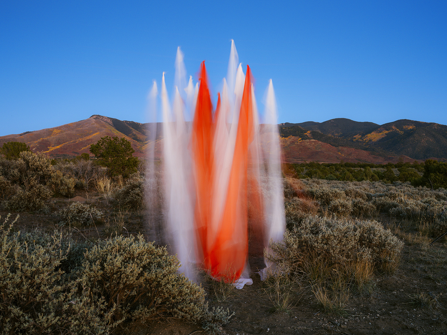 A dramatic fountain of white water and bright red silk erupts from the desert floor, surrounded by sparse vegetation and distant mountains under a clear blue sky.