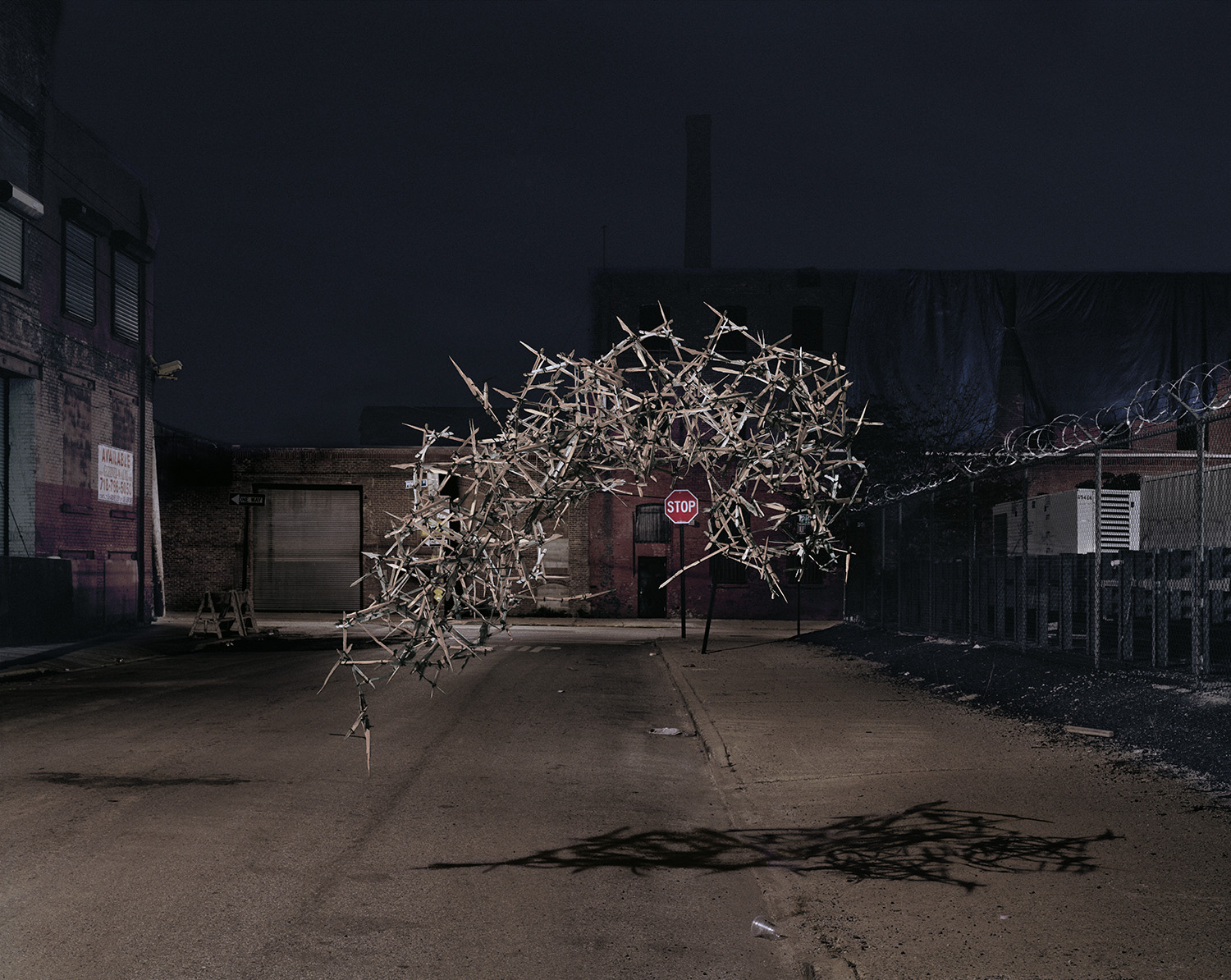 Nighttime photograph of an industrial urban courtyard featuring a skeletal wooden structure illuminated against dark brick buildings and chain-link fencing. The fragmented, branch-like form casts sharp shadows on the pavement below, positioned near a red stop sign.