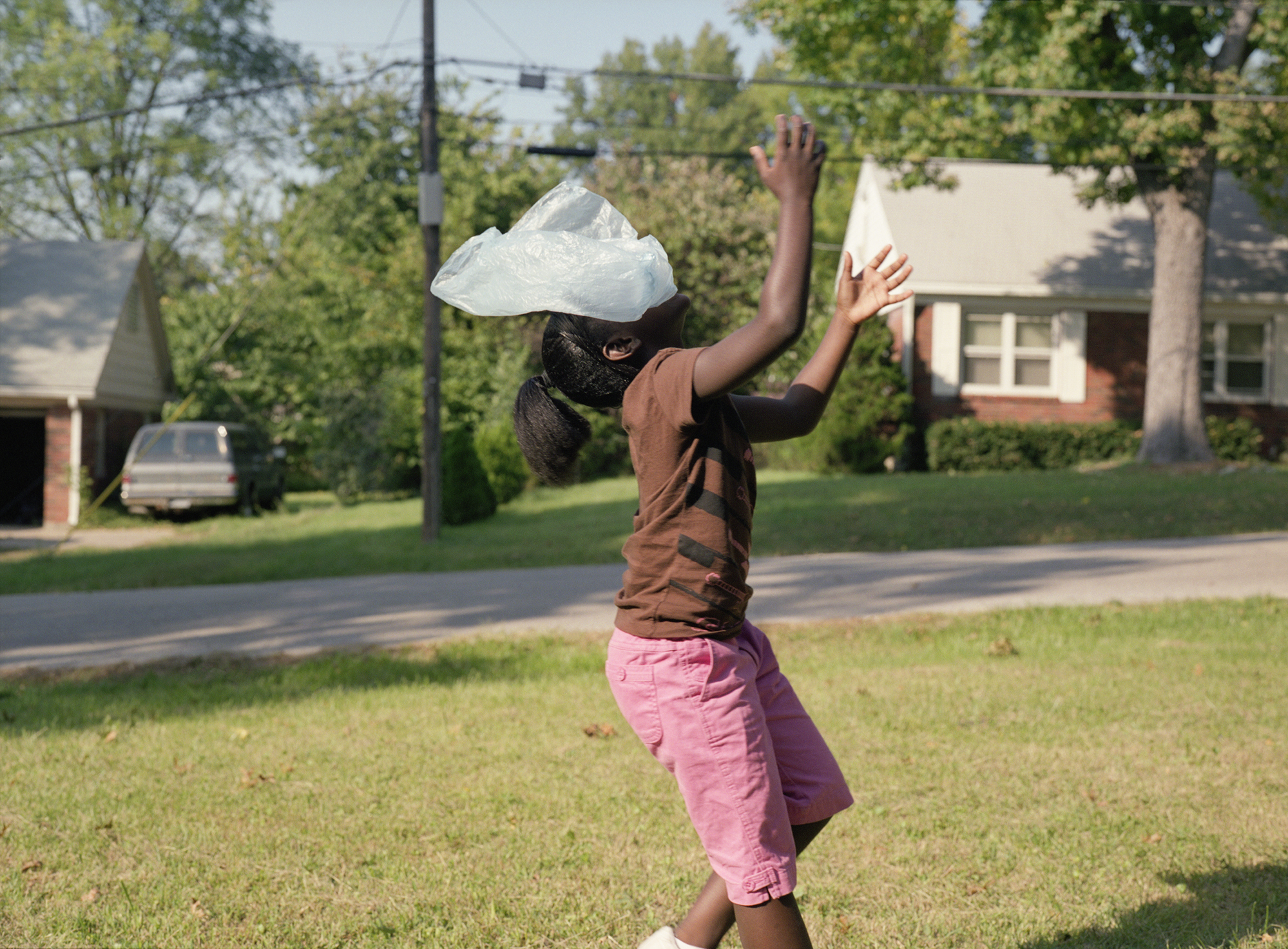 A figure stands in a residential street wearing a brown shirt and pink pants, with their head completely covered by a translucent white plastic bag. Their arms are raised expressively against a backdrop of tree-lined houses and suburban architecture.