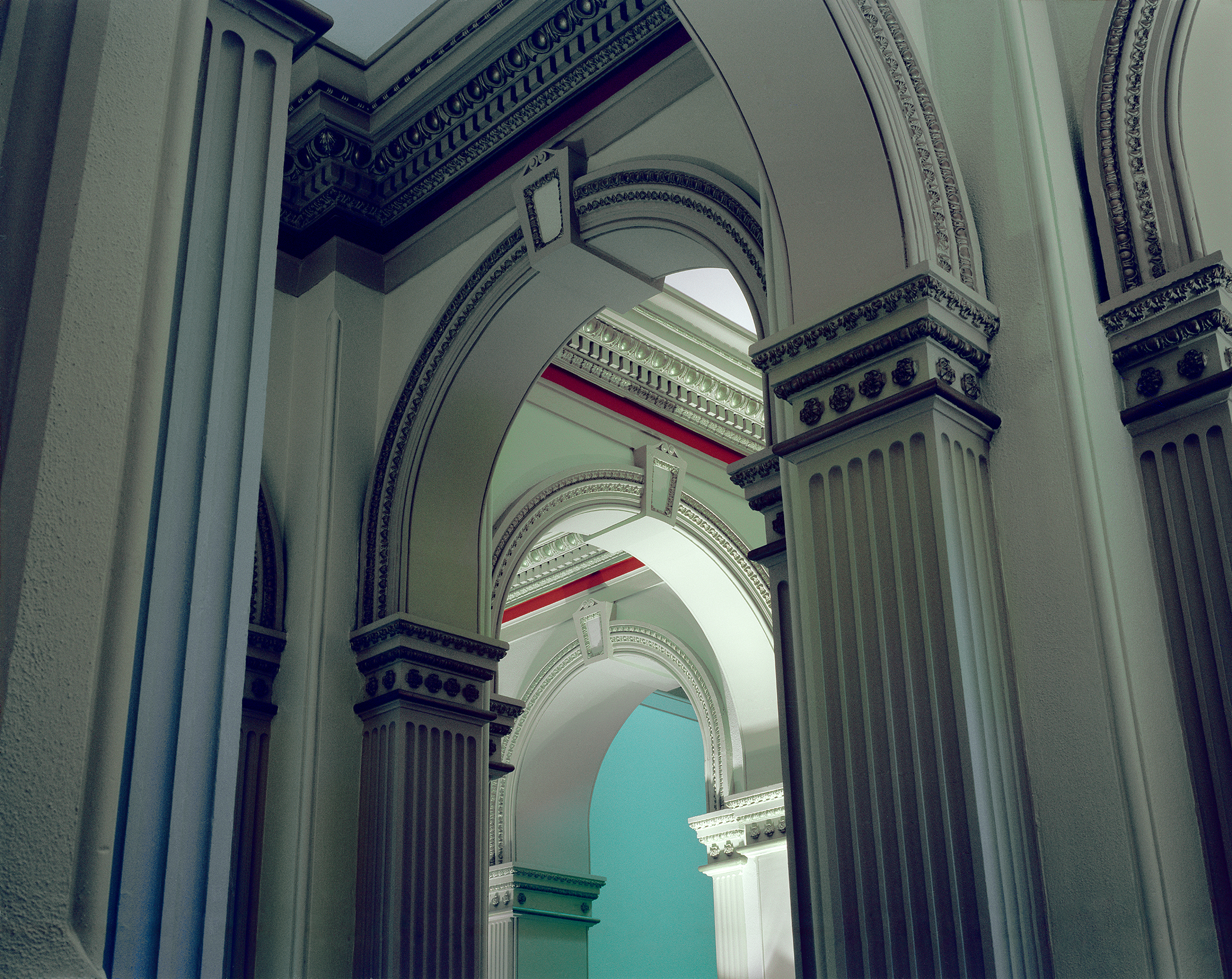 A symmetrical architectural corridor receding into depth, featuring classical arched doorways with ornate moldings and fluted columns in white and dark blue. Glimpses of colored walls—turquoise, red, and green—visible through successive archways, creating a layered perspective down the hallway.