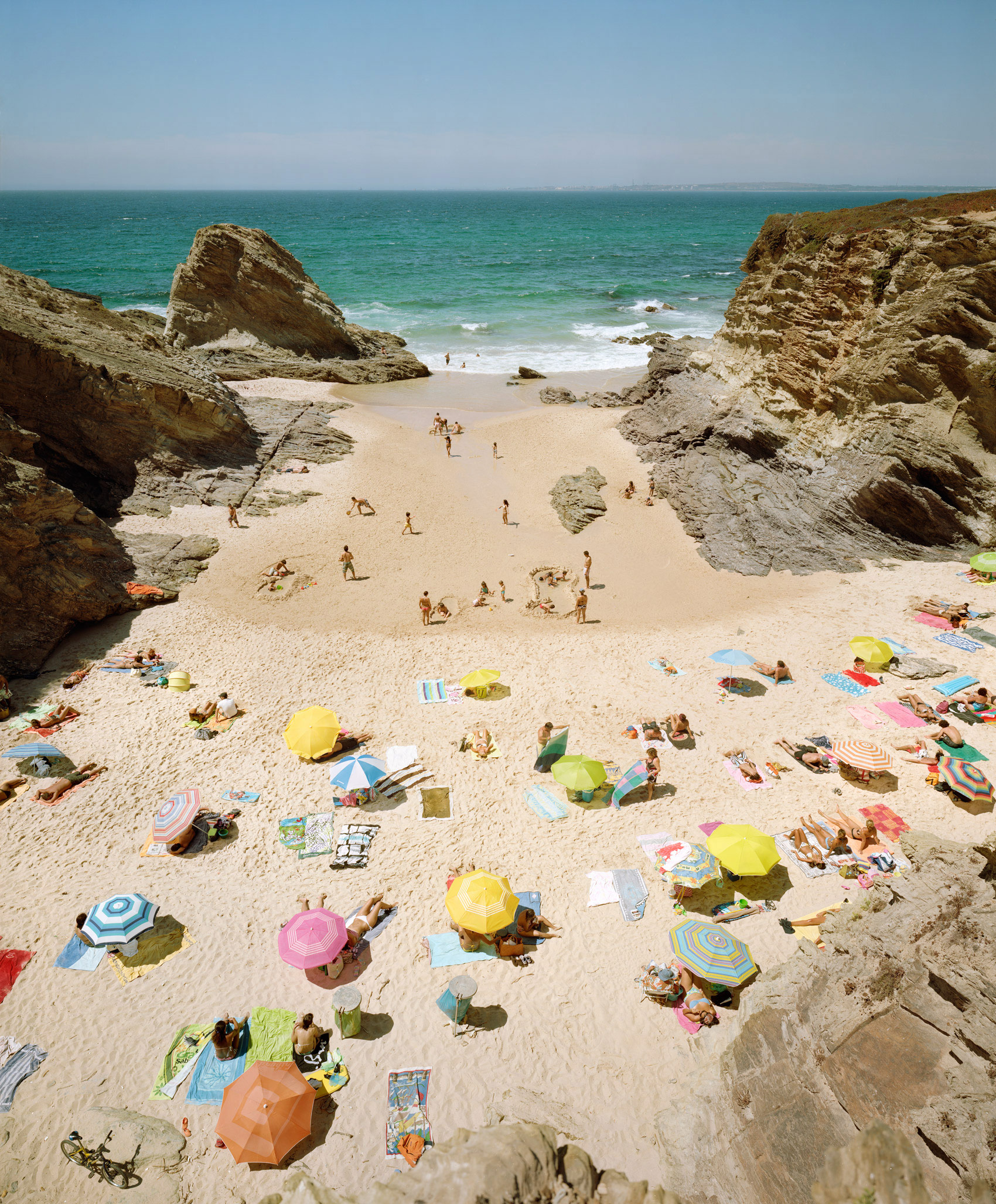 Aerial view of a sandy beach cove flanked by towering golden rock formations, dotted with colorful umbrellas and sunbathers scattered across the sand, with turquoise ocean and white waves beyond.