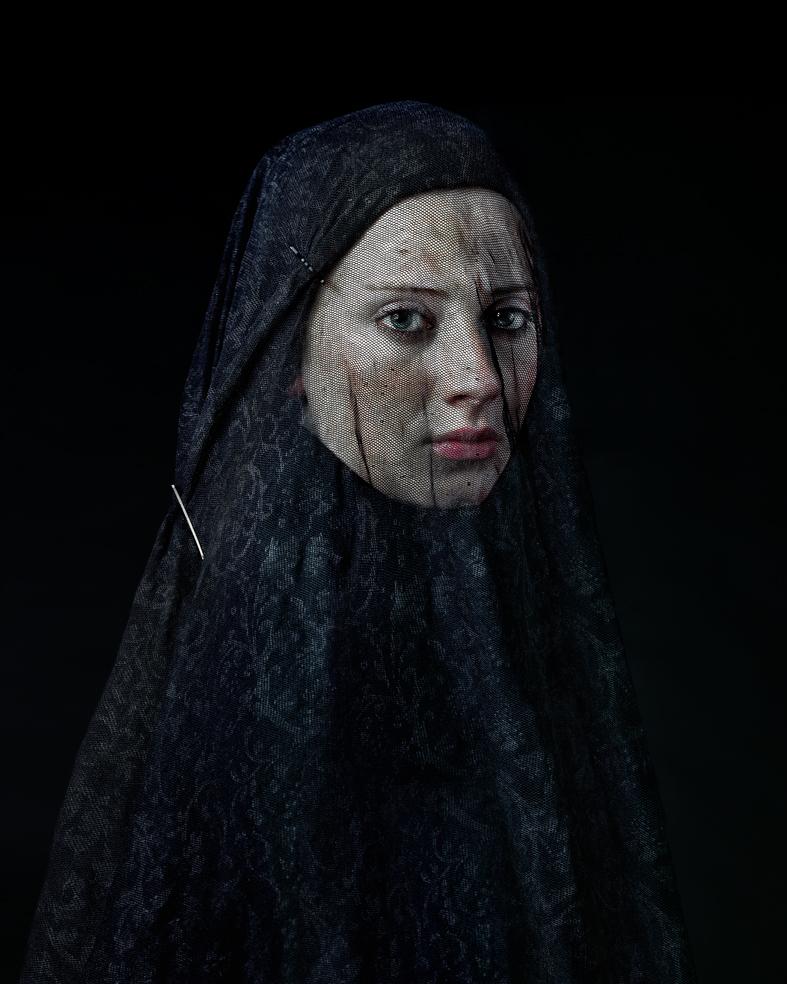 Portrait of a person wearing a dark, textured head covering against a black background. The figure gazes directly toward the camera with a composed expression, their pale face contrasting sharply with the deep navy fabric that drapes over their head and shoulders.