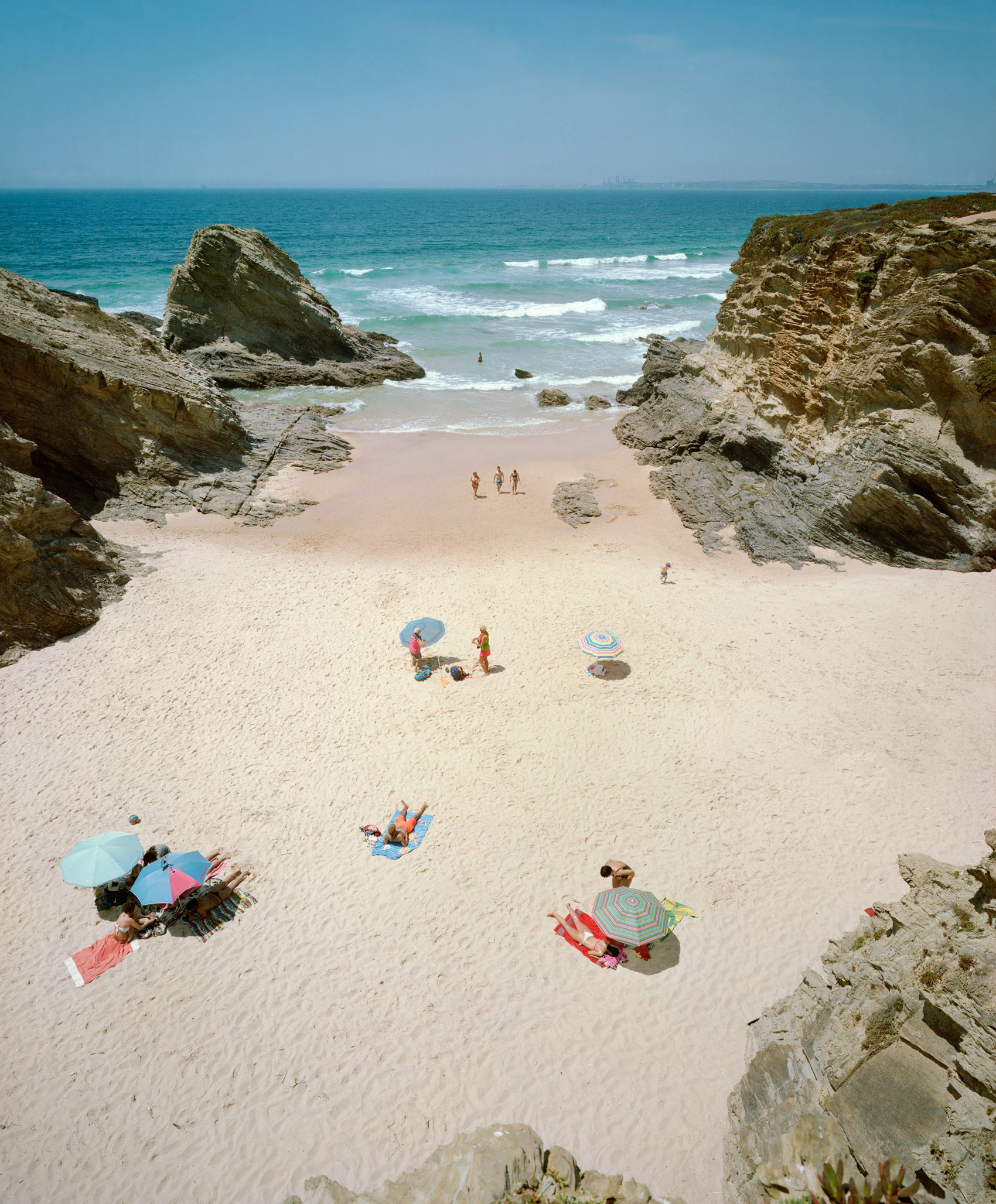 Aerial view of a secluded beach cove framed by dramatic rocky outcrops, with scattered beachgoers beneath colorful umbrellas and on towels across pale sand, turquoise waves visible beyond.