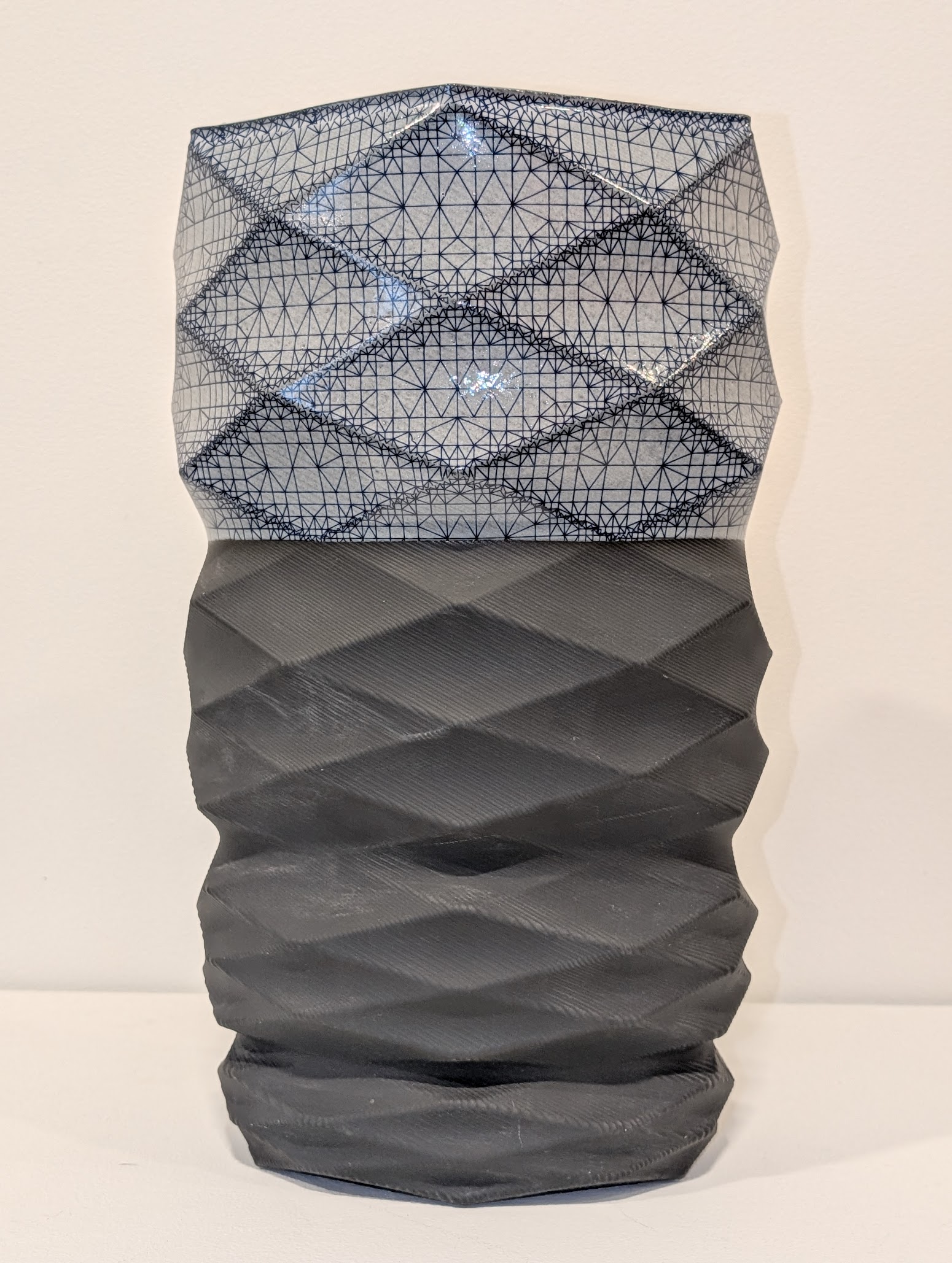 A cylindrical sculptural form composed of two distinct sections: an upper portion of delicate wire mesh with geometric patterning in shades of blue and gray, and a lower section of solid dark gray material with organic, folded surface textures. The piece stands vertically against a neutral background.