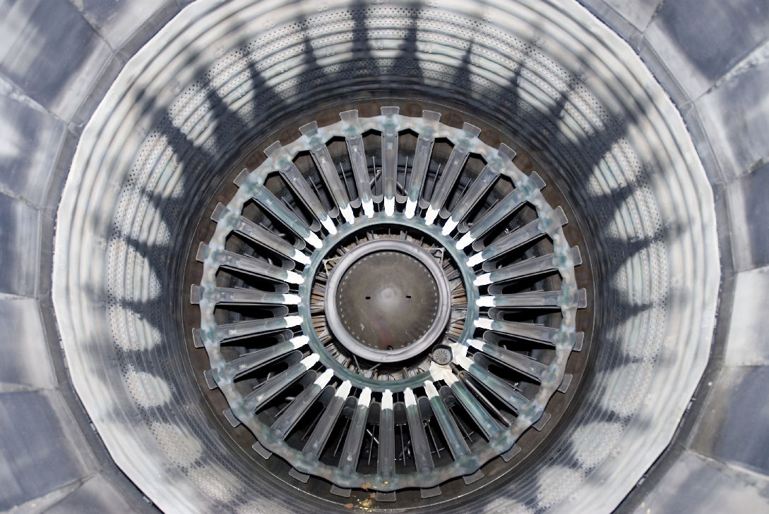 Concentric circular view down the interior of a jet engine, featuring radiating metal blades arranged in perfect symmetry around a central dome, with layered rings of perforated metal creating a sense of deep perspective and industrial precision.