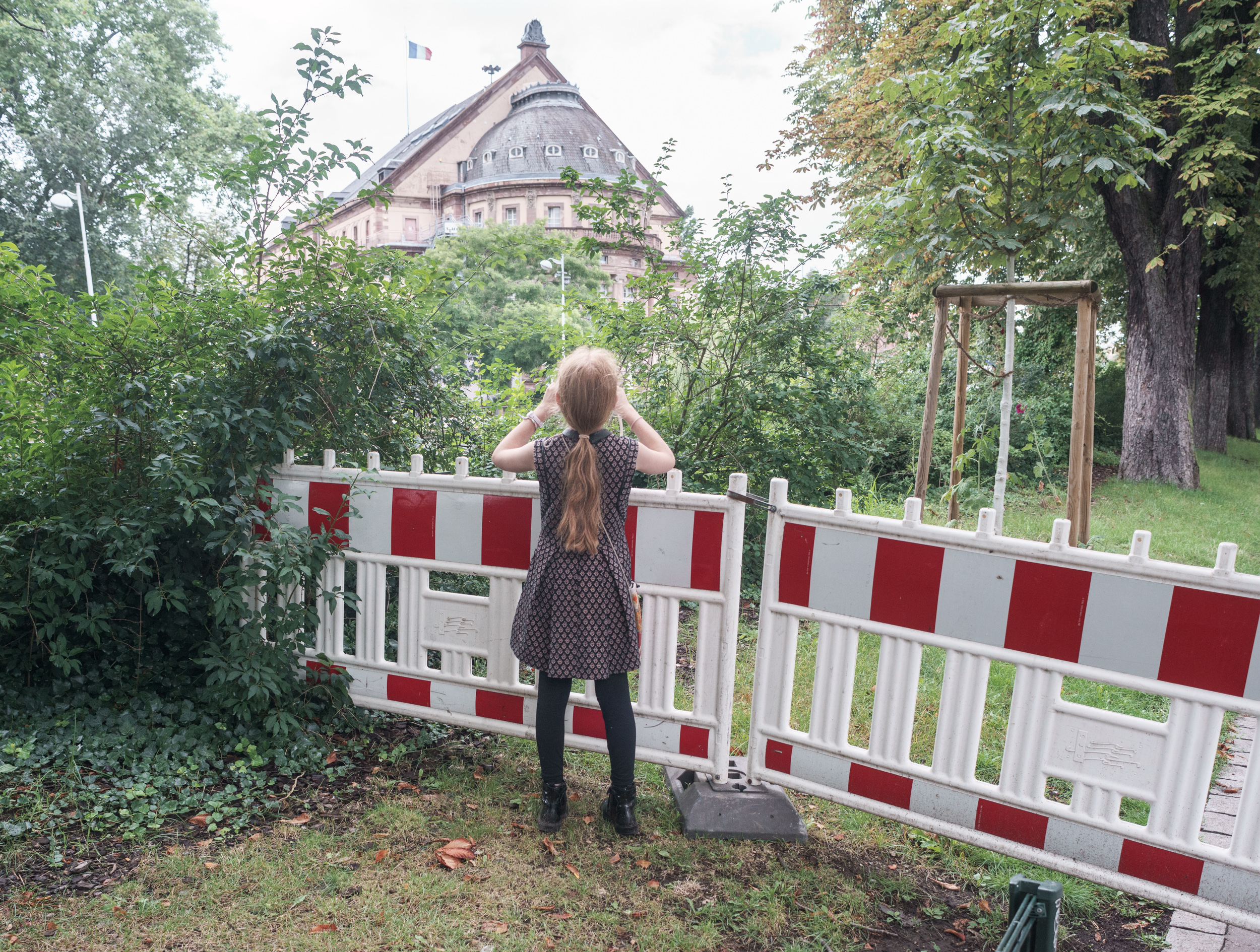 A young girl with long hair stands facing away from the camera at a red and white striped barrier in a park setting, her hands raised to her head. Behind her, a distinctive pink neoclassical building with a domed roof and French flag is visible through the trees, creating a striking contrast between the industrial barrier and the architectural landmark.