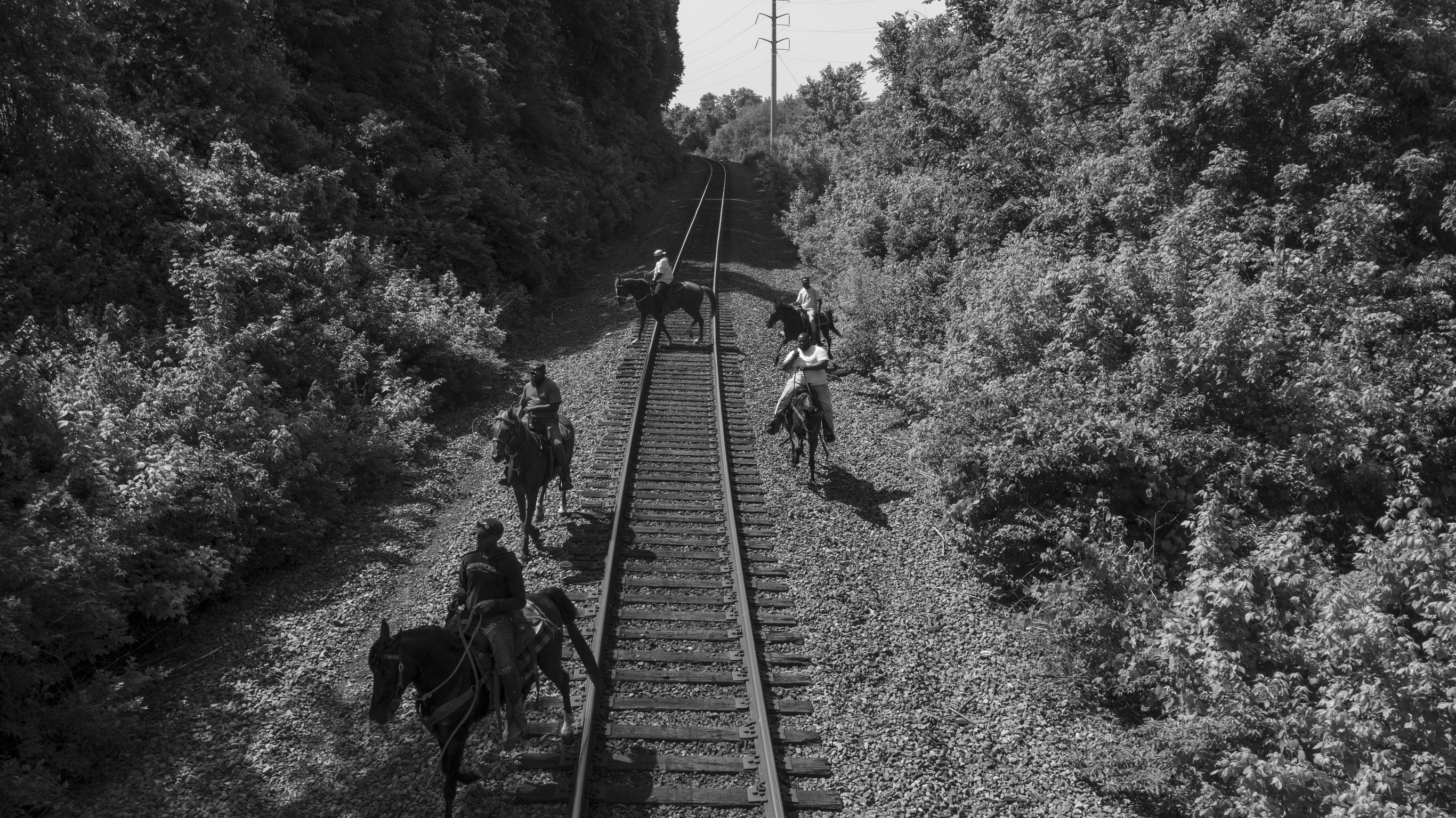 Black and white photograph showing five mounted horsemen positioned on and alongside railroad tracks that converge toward the horizon, flanked by dense vegetation on both sides. The composition uses linear perspective to create depth, with a utility pole visible in the distant sky.