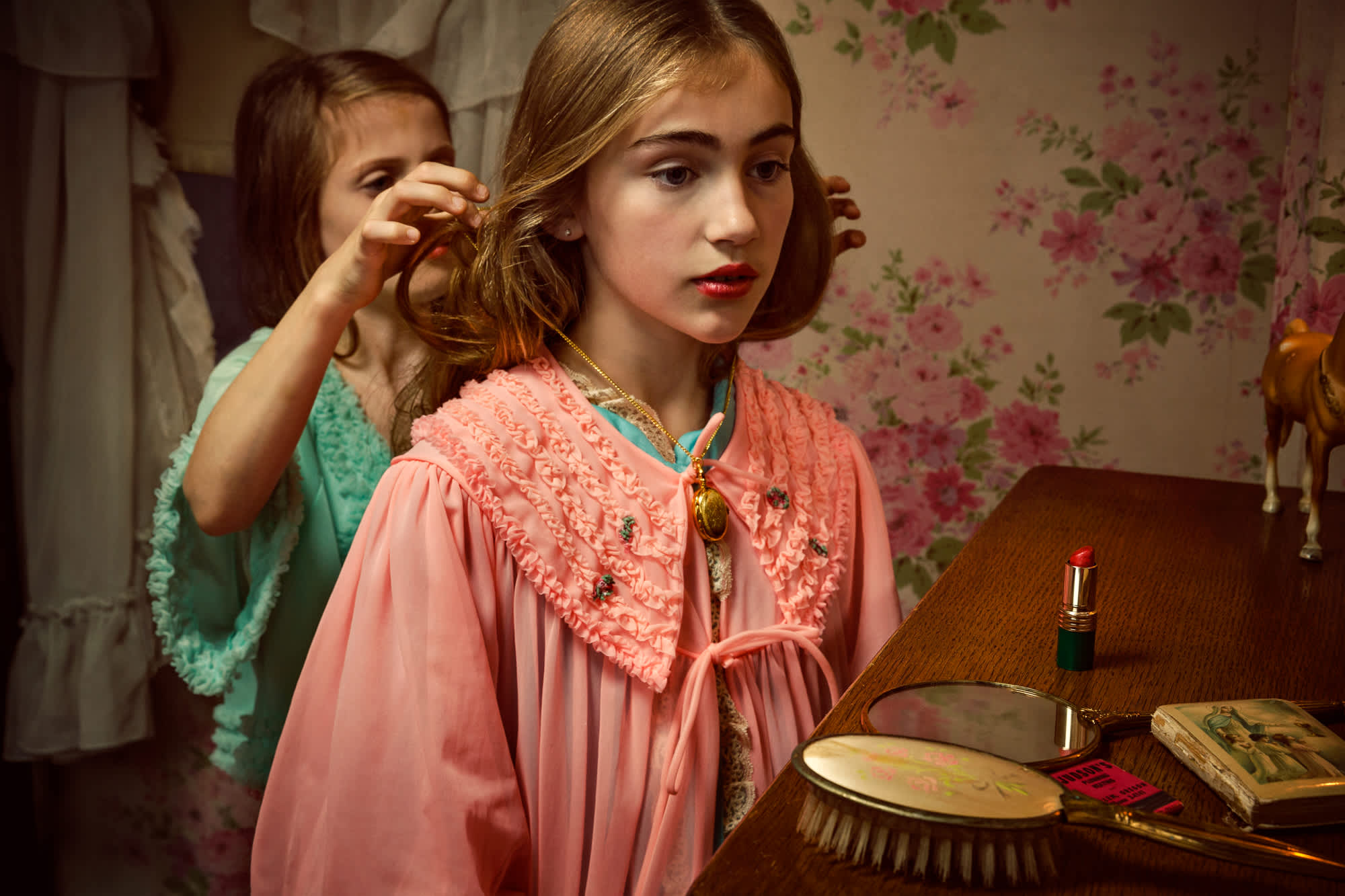 Two young girls in vintage-inspired pink and mint green clothing stand at a wooden vanity in an interior with floral wallpaper. The girl in focus, wearing a pink robe with an ornate ruched collar, gazes downward while adjusting her hair; behind her, a second girl in green watches. The vanity holds a brush, mirror, lipstick, and open books, rendered in warm, naturalistic tones with careful attention to period detail and intimate domestic atmosphere.
