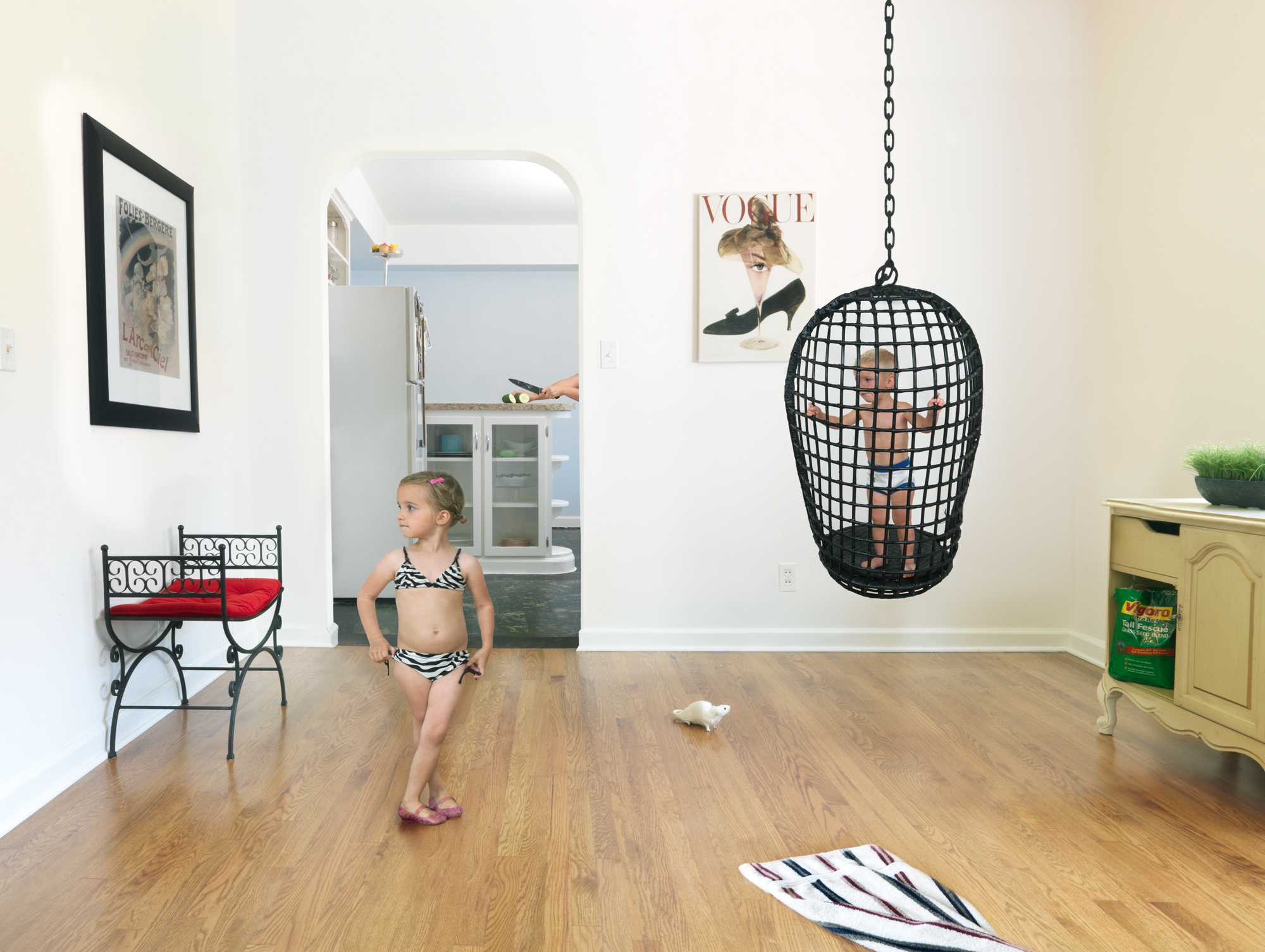 A young girl in a striped swimsuit stands on a wooden floor in a bright, domestic interior space, glancing toward a black wicker hanging chair suspended from the ceiling. The room contains vintage-styled furnishings including a decorative metal bench with red cushioning, framed artwork on white walls, and a yellow cabinet, creating a carefully composed scene that blends domestic comfort with surreal sculptural elements.