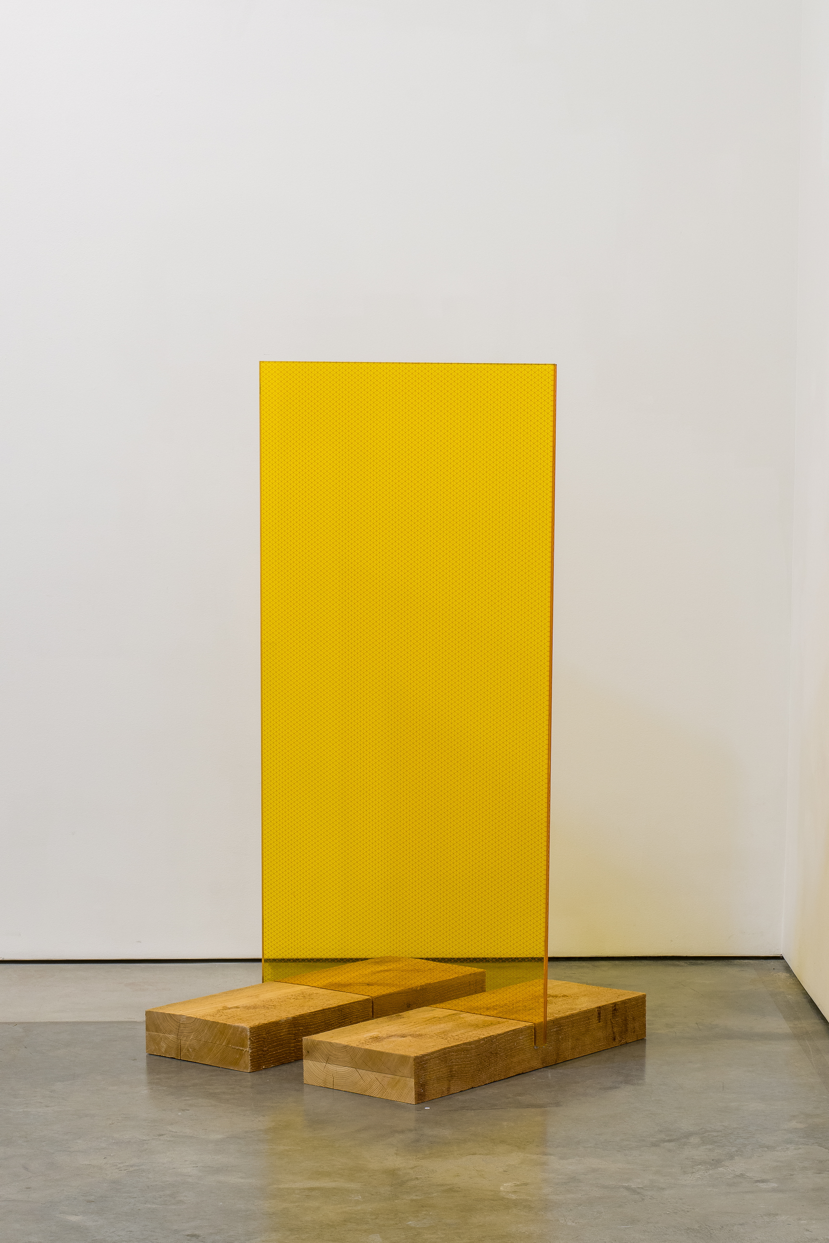 A bright yellow rectangular panel stands vertically on a polished concrete floor, supported by two natural wood blocks positioned beneath it. The panel's textured surface catches light, creating subtle variations in tone, while the wooden base elements are left raw and unfinished, creating a stark material contrast in a minimalist gallery setting.
