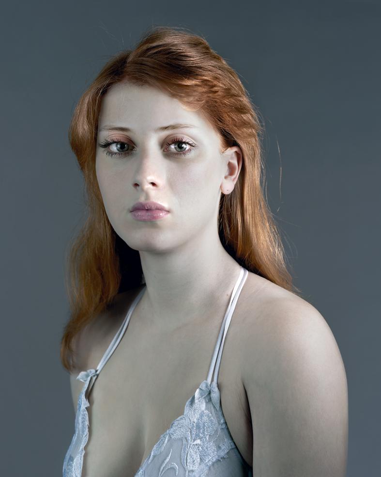 Portrait of a woman with long reddish-brown hair gazing downward, wearing a light blue lace halter top against a neutral gray background. The photograph employs soft, directional lighting that emphasizes the subject's pale complexion and contemplative expression.