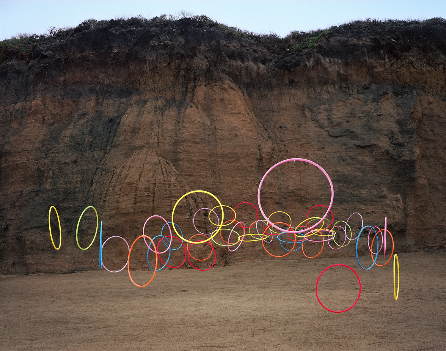 Colorful hula hoops in various sizes and hues—yellow, pink, red, blue, and orange—are arranged in an overlapping horizontal line across a sandy beach, set against a tall cliff face with dark soil and sparse vegetation at its top.