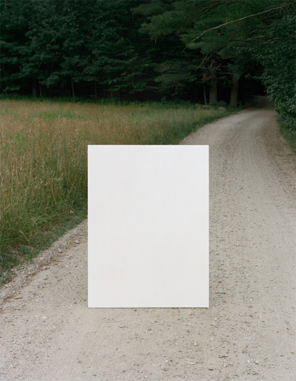 A white rectangular panel stands vertically on a gravel road lined with overgrown grass and dense green trees, creating a stark contrast between the blank white form and the natural landscape behind it.