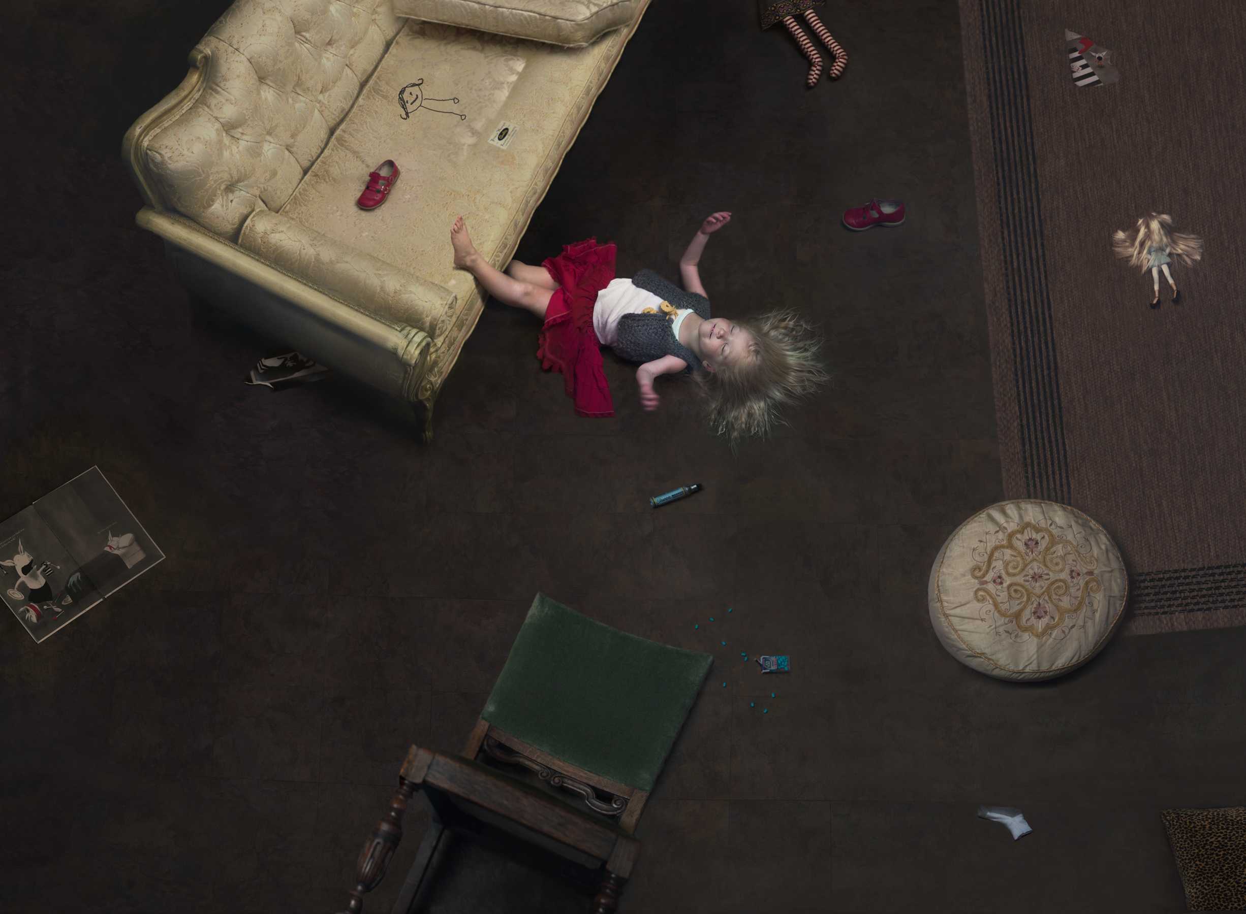 Overhead view of a young child lying on a dark floor surrounded by scattered toys, shoes, and household items, with a cream-colored upholstered sofa visible in the upper left corner. The child wears a red garment and white socks against the stark contrast of the dark flooring.