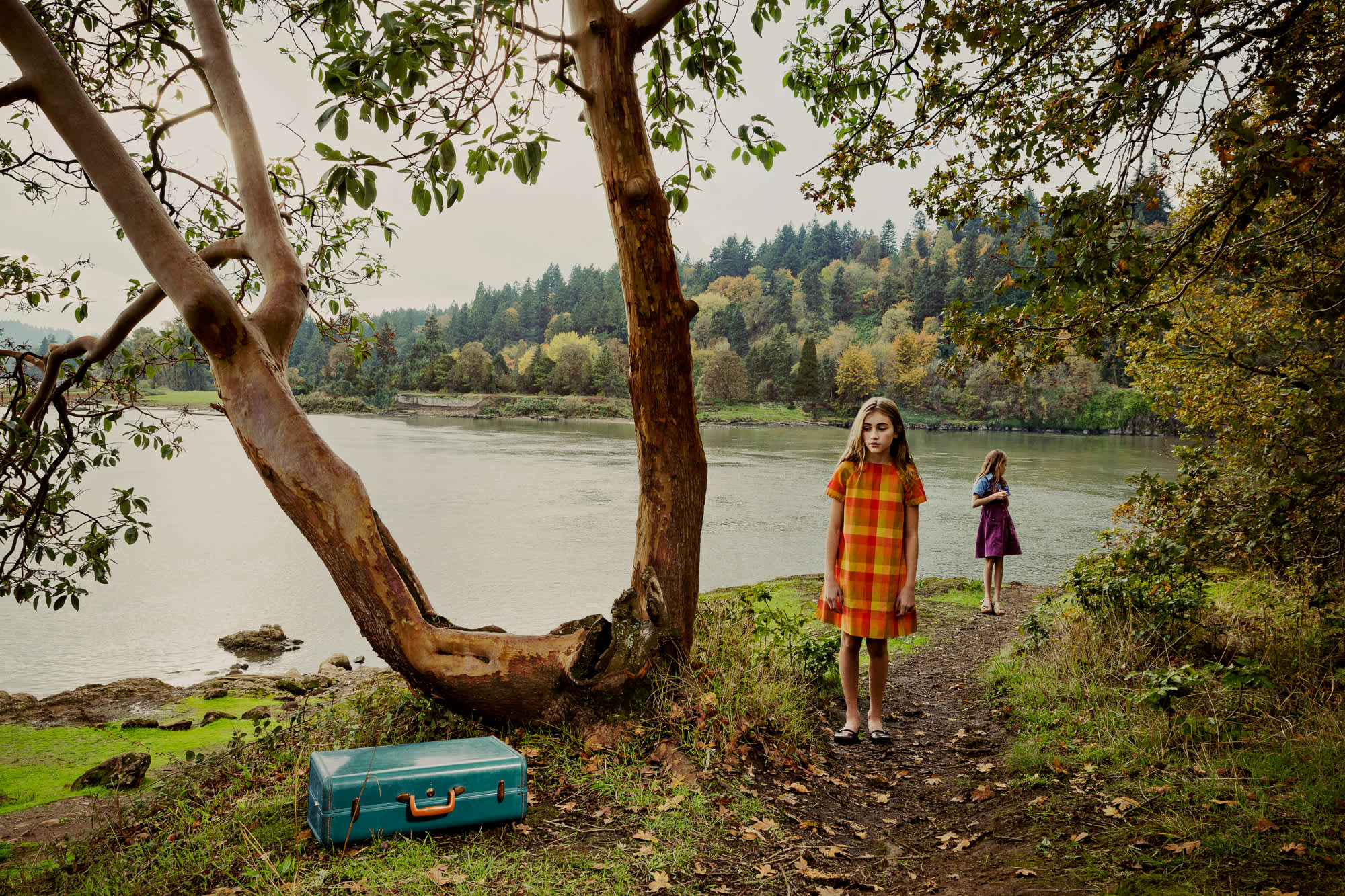 Two young girls stand on a wooded riverbank beneath gnarled trees with exposed roots, one wearing a red and yellow plaid dress in the foreground and another in a purple dress further back. A turquoise vintage suitcase rests on the mossy ground in the lower left, while a calm river and forested hillside stretch across the background under an overcast sky.