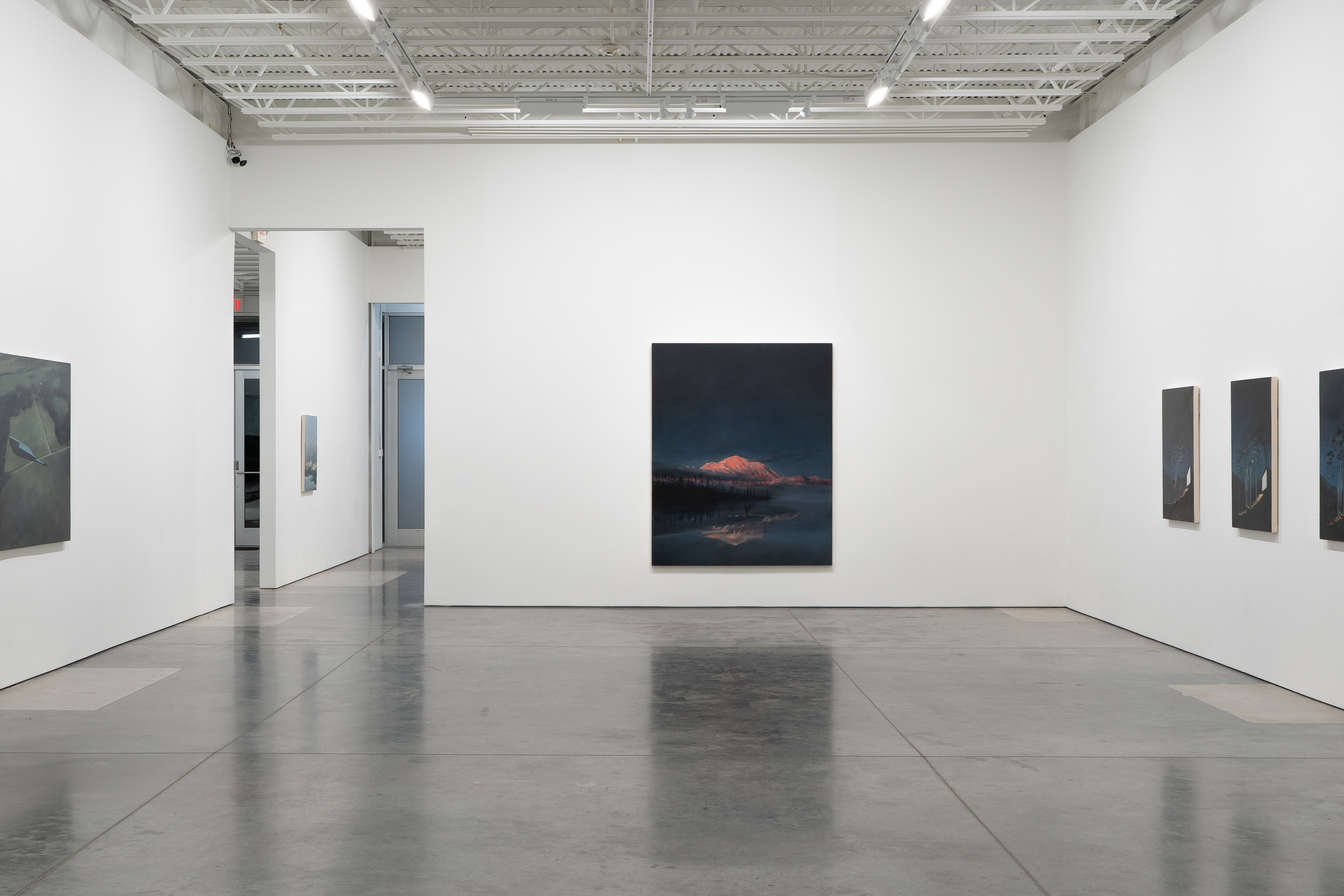 A minimalist gallery interior with polished concrete floors and white walls displays several paintings. The central work depicts a snow-capped mountain glowing red against a dark night sky, reflected in still water below. Additional smaller dark paintings are positioned on the right wall, while doorways and skylights punctuate the left side of the space.
