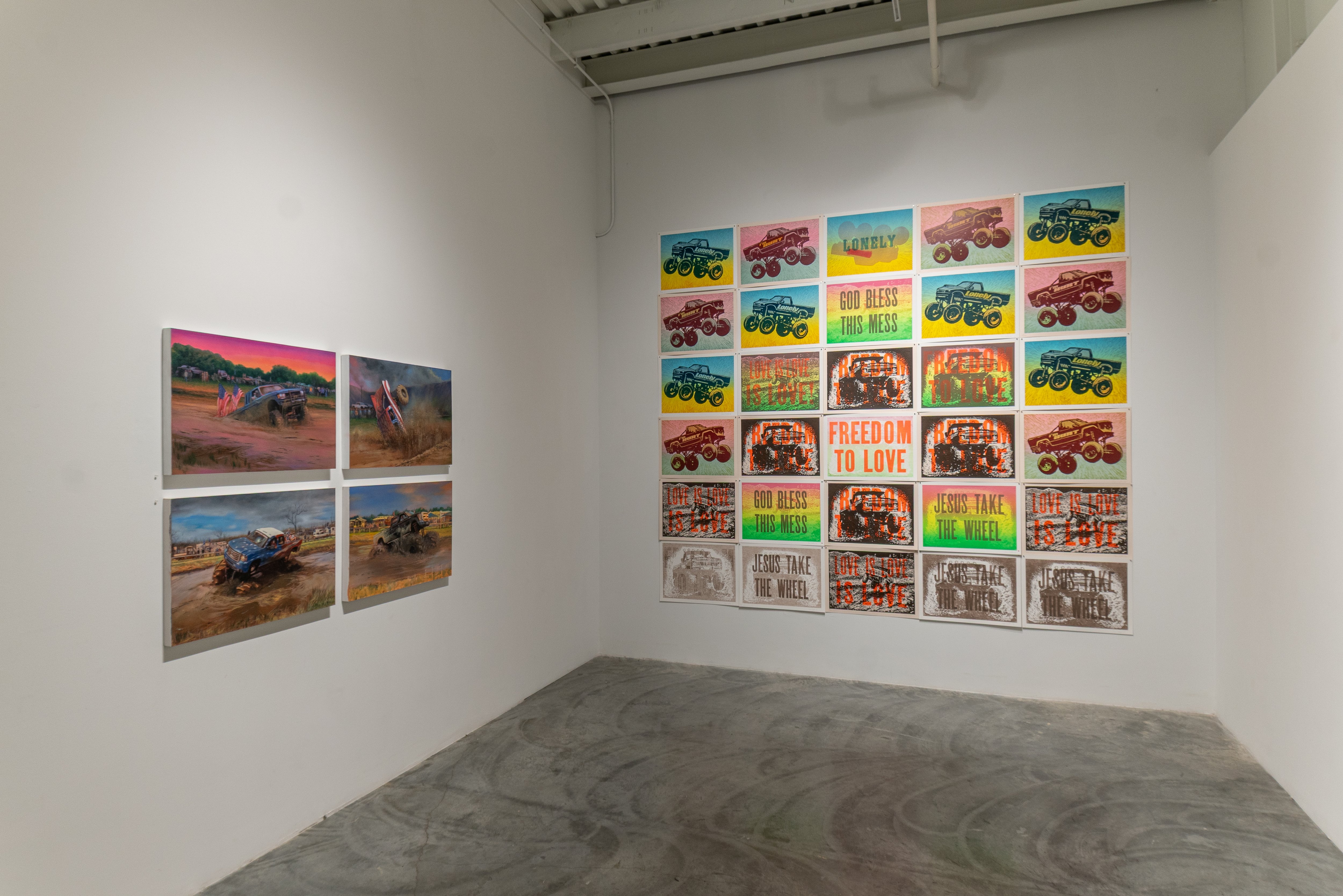 Gallery installation view showing four large photographic prints of monster trucks in desert landscapes mounted on the left wall, and a grid of approximately twenty smaller colorful prints featuring monster trucks with overlaid text phrases including "God Bless This Mess," "Freedom to Love," and "Jesus Take the Wheel" mounted on the right wall; concrete floor visible in foreground.