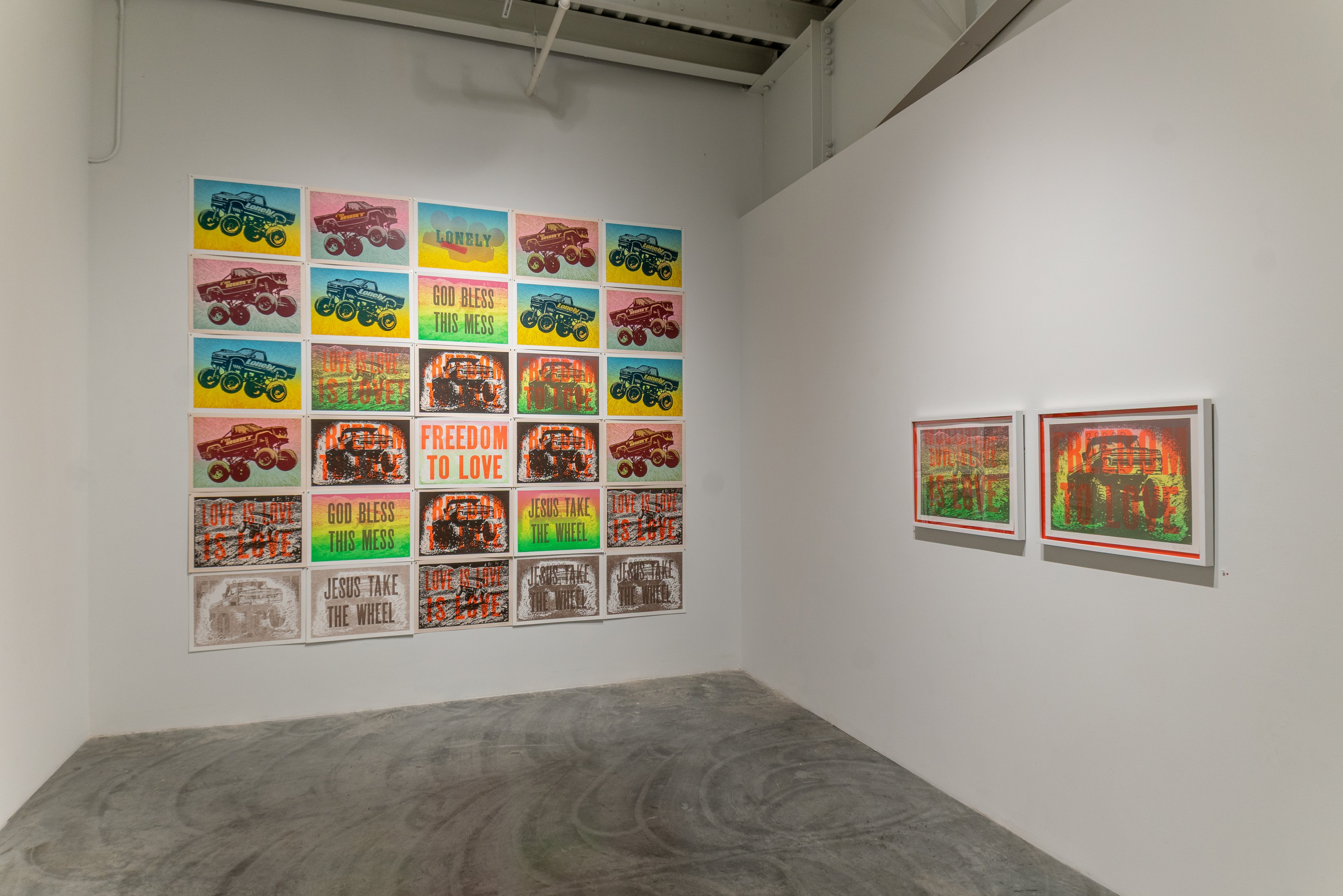A gallery corner displays a large grid of colorful prints featuring monster trucks in various compositions on the left wall, interspersed with bright text passages in yellow, green, and pink including phrases like "God Bless This Mess" and "Freedom to Love." The right wall shows two framed prints with similar monster truck imagery and warm orange-red tones. The concrete floor is marked with circular patterns.