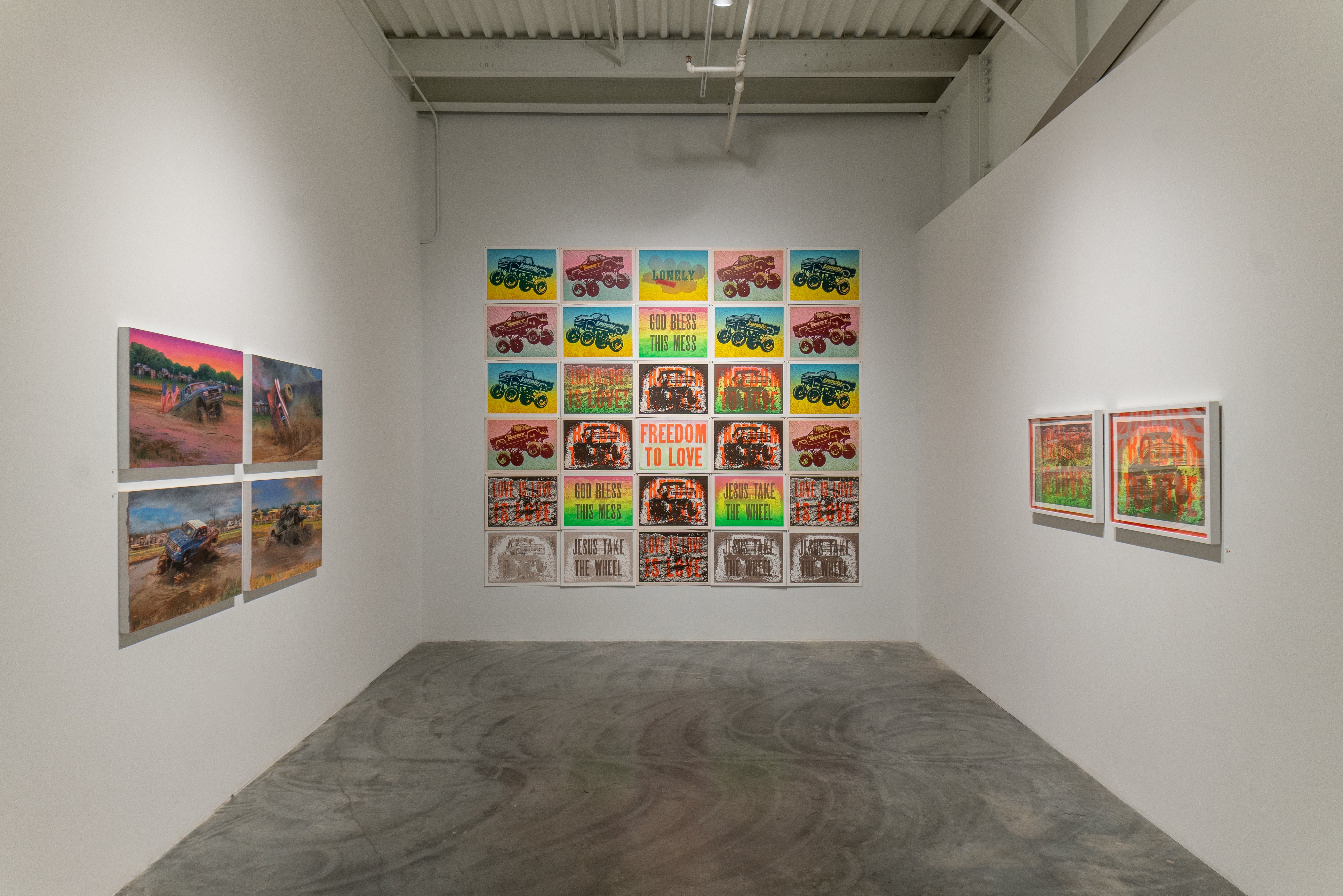 Gallery installation view featuring four photographs of monster trucks in desert landscapes mounted on the left and right walls, and a large grid of colorful pop art-style prints depicting monster trucks with neon text phrases including "LONELY," "GOD BLESS THIS MESS," "FREEDOM TO LOVE," and "JESUS TAKE THE WHEEL" mounted on the back wall.