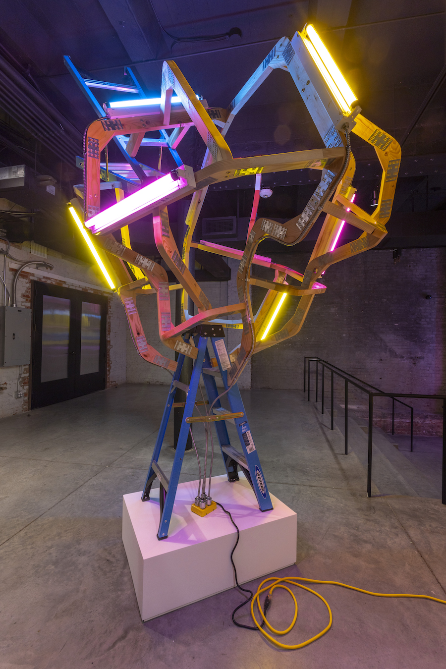 Colorful neon light installation featuring wooden ladders and structural beams arranged vertically on a pink platform. Bright blue, magenta, and yellow fluorescent tubes intersect across the composition, creating an energetic geometric form against the gallery's industrial brick walls.
