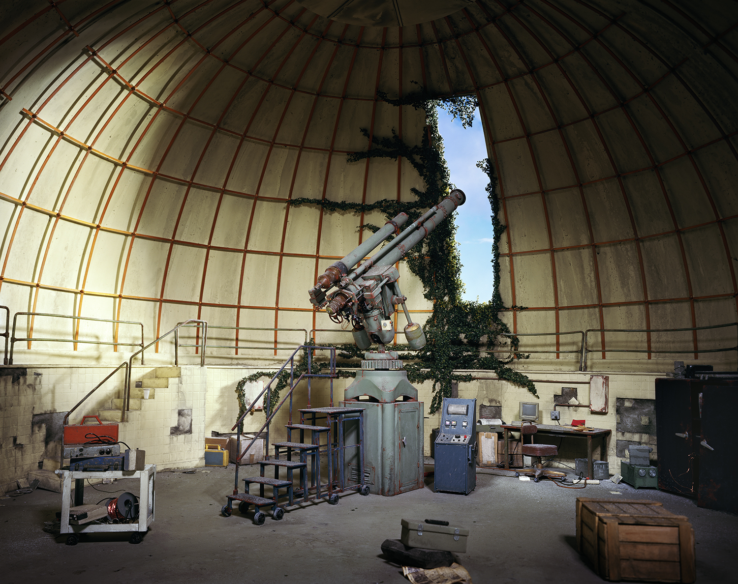 A large brass telescope mounted on a pedestal stands centered within an abandoned observatory dome, surrounded by weathered equipment, control panels, and scattered debris. Ivy creeps across the dome's interior walls and frames the opening above, while natural light streams in from a bright blue sky visible through the dome's aperture.