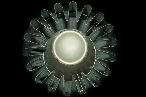 A spherical form with a luminous cream-colored center is surrounded by a radial arrangement of dark metallic or glass tube-like structures that fan outward symmetrically against a black background, creating a geometric, almost mechanical flower-like composition.