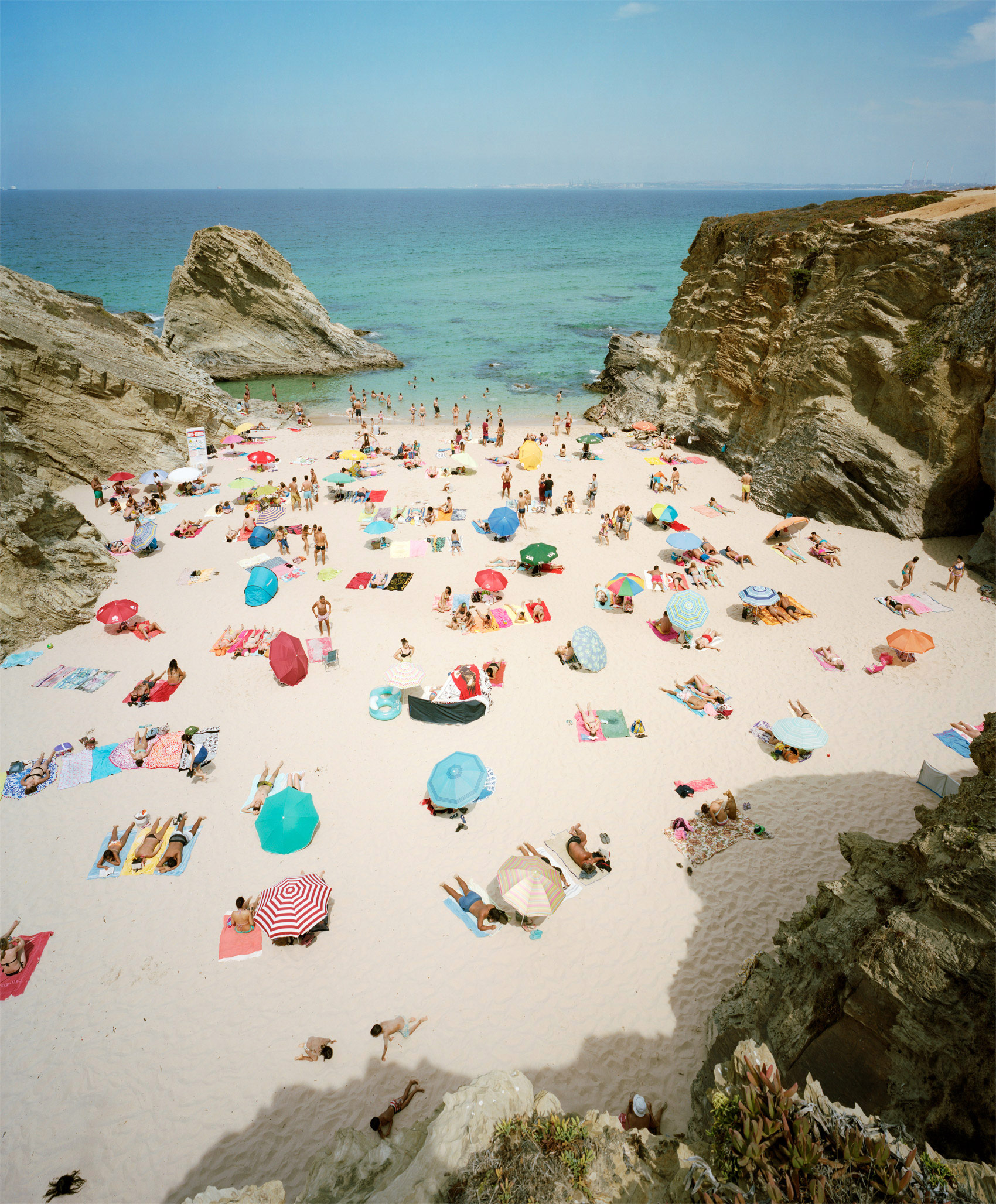 Aerial view of a crowded beach nestled between dramatic limestone cliffs, with numerous colorful umbrellas and sunbathers scattered across white sand, turquoise water visible beyond.