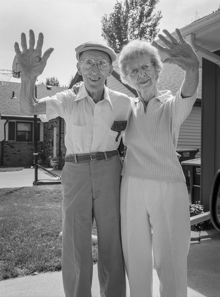 Black and white photograph of an elderly man and woman standing in a residential driveway, both waving at the camera with their arms raised. The man wears glasses, a cap, and a light-colored shirt; the woman wears a ribbed sweater with white hair. Houses and trees are visible in the background.