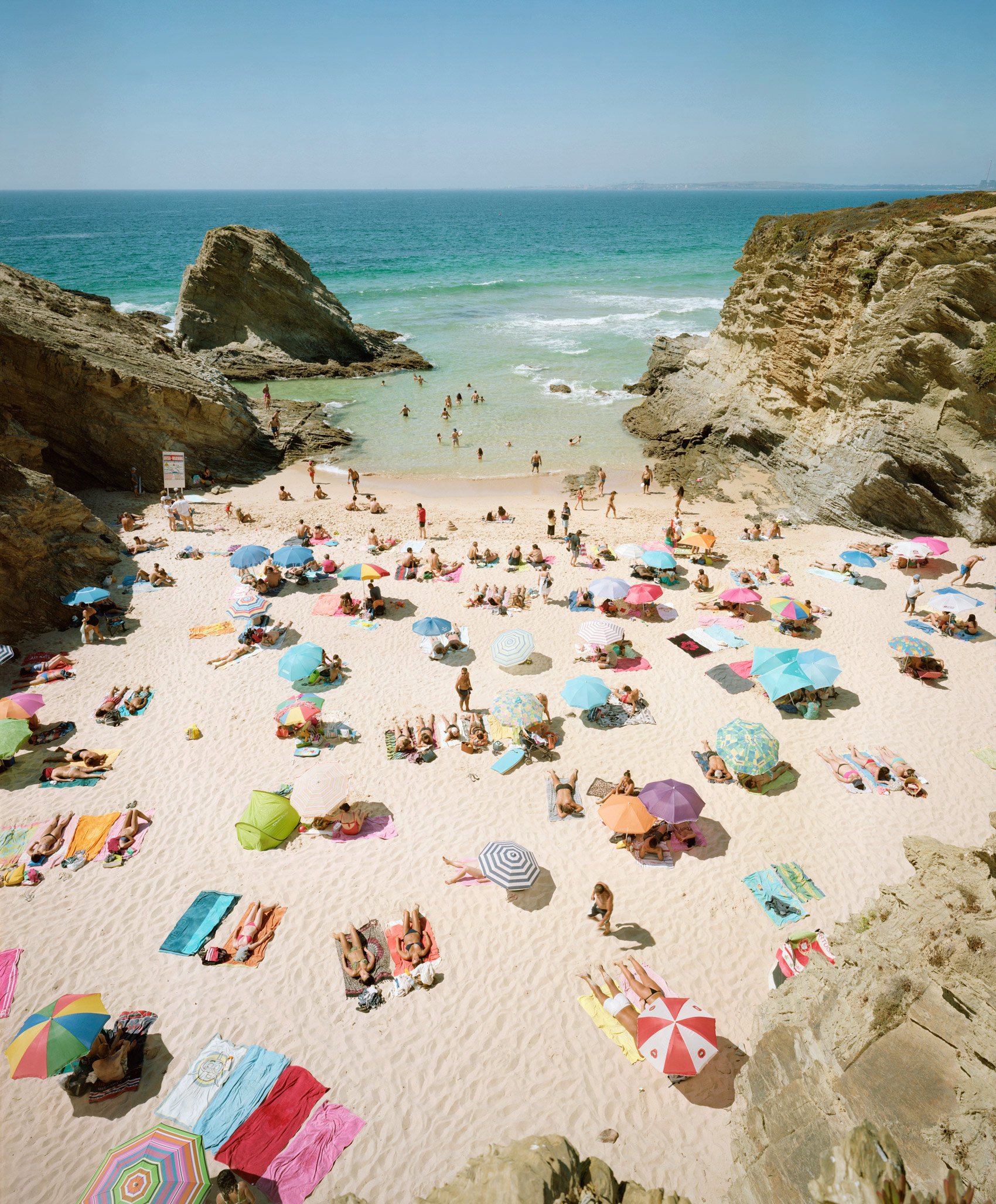 Aerial view of a crowded sandy beach nestled between dramatic limestone cliffs, with numerous colorful umbrellas and beach towels scattered across the sand, beachgoers swimming and wading in turquoise waters, and calm blue sky above.