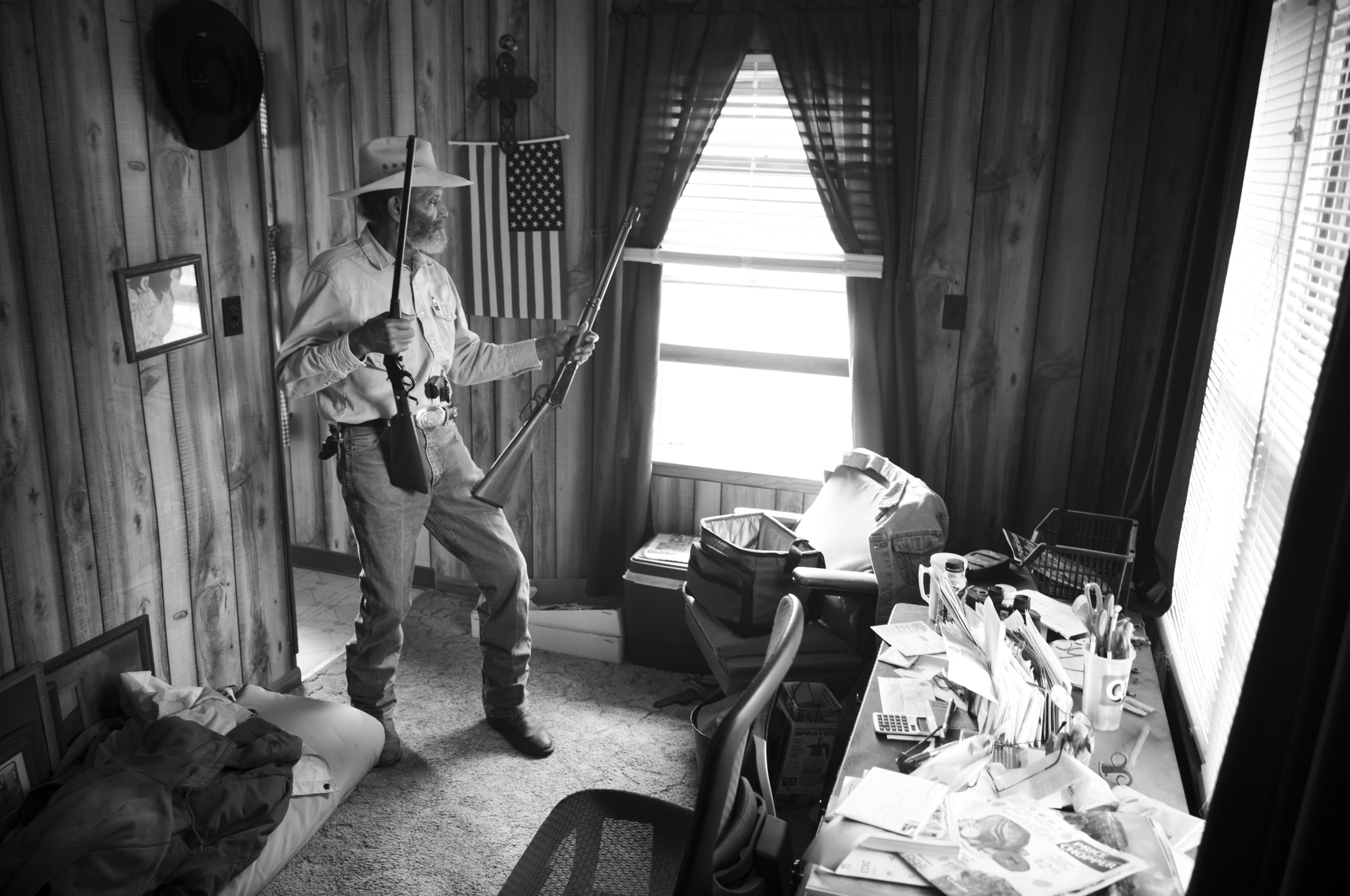Black and white photograph of a man in a cowboy hat and period clothing posed in a modest interior space, holding a rifle and an American flag. The room features wooden walls, a bright window with curtains, and a cluttered desk scattered with papers and supplies, creating a stark contrast between the figure's dramatic stance and the everyday domestic setting.