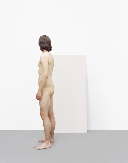 Nude figure viewed from the side standing against a white rectangular panel in a minimalist gallery space, rendered in muted flesh tones and grays with soft, diffused lighting.
