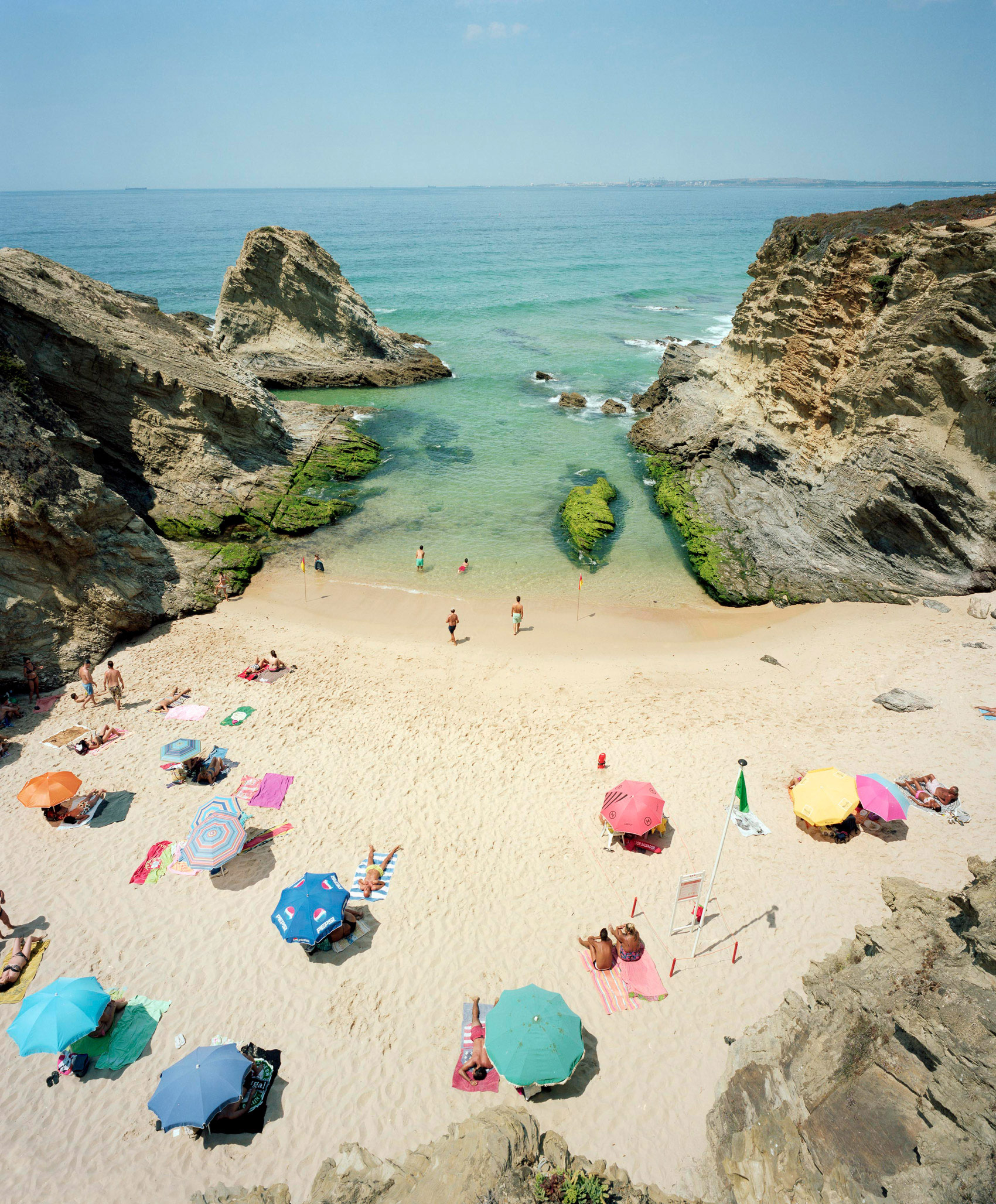 Aerial view of a crowded beach cove enclosed by dramatic limestone cliffs, with turquoise water and white sand dotted with colorful beach umbrellas and sunbathers. Swimmers wade in the shallow water while green algae-covered rocks create natural divisions in the bay.