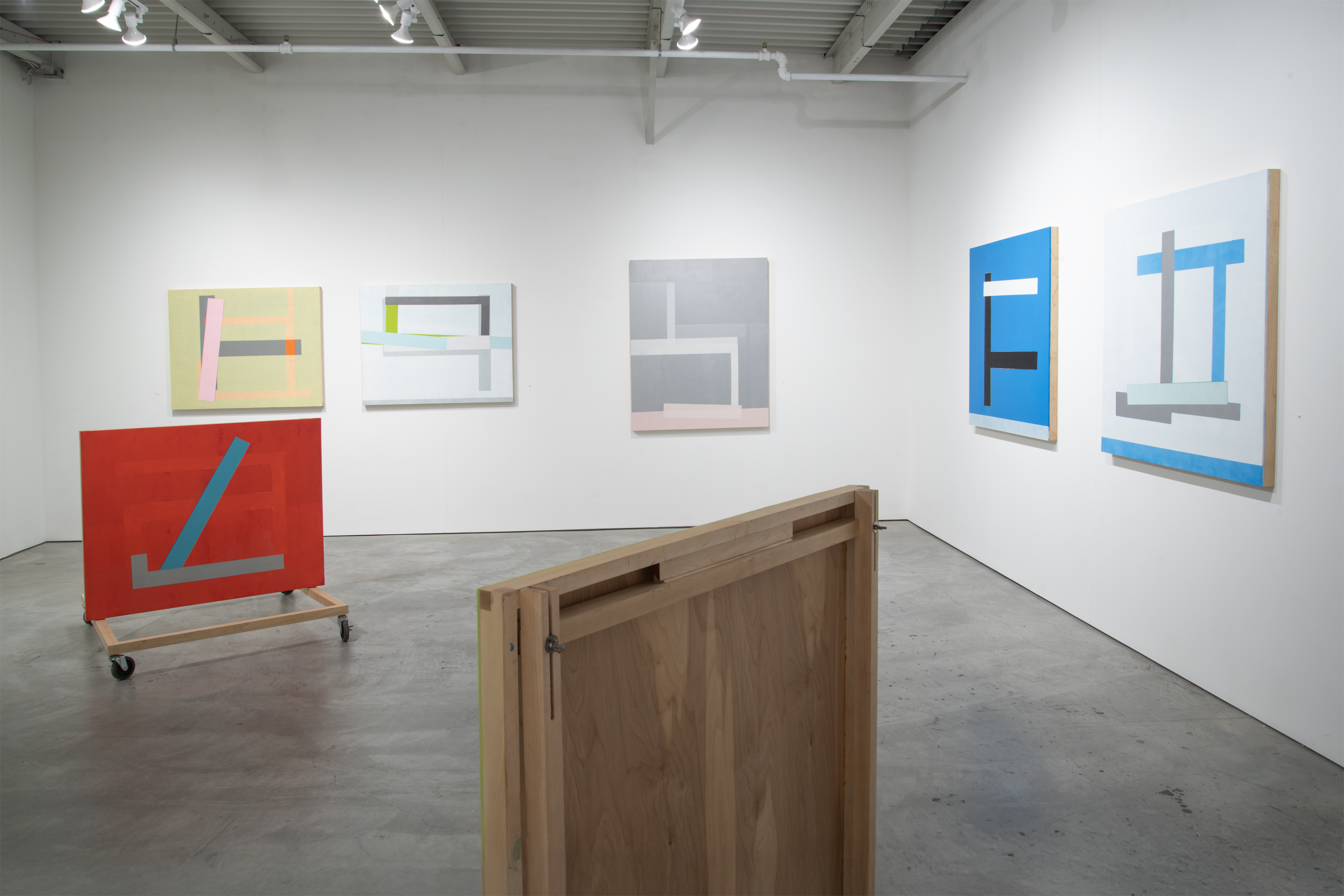 Contemporary gallery installation featuring five abstract paintings with geometric forms in muted and bold colors—pastels, grays, blues, and red—arranged on white walls, alongside two wooden sculptural elements on the concrete floor, creating a unified spatial composition exploring formal relationships between color, form, and environment.