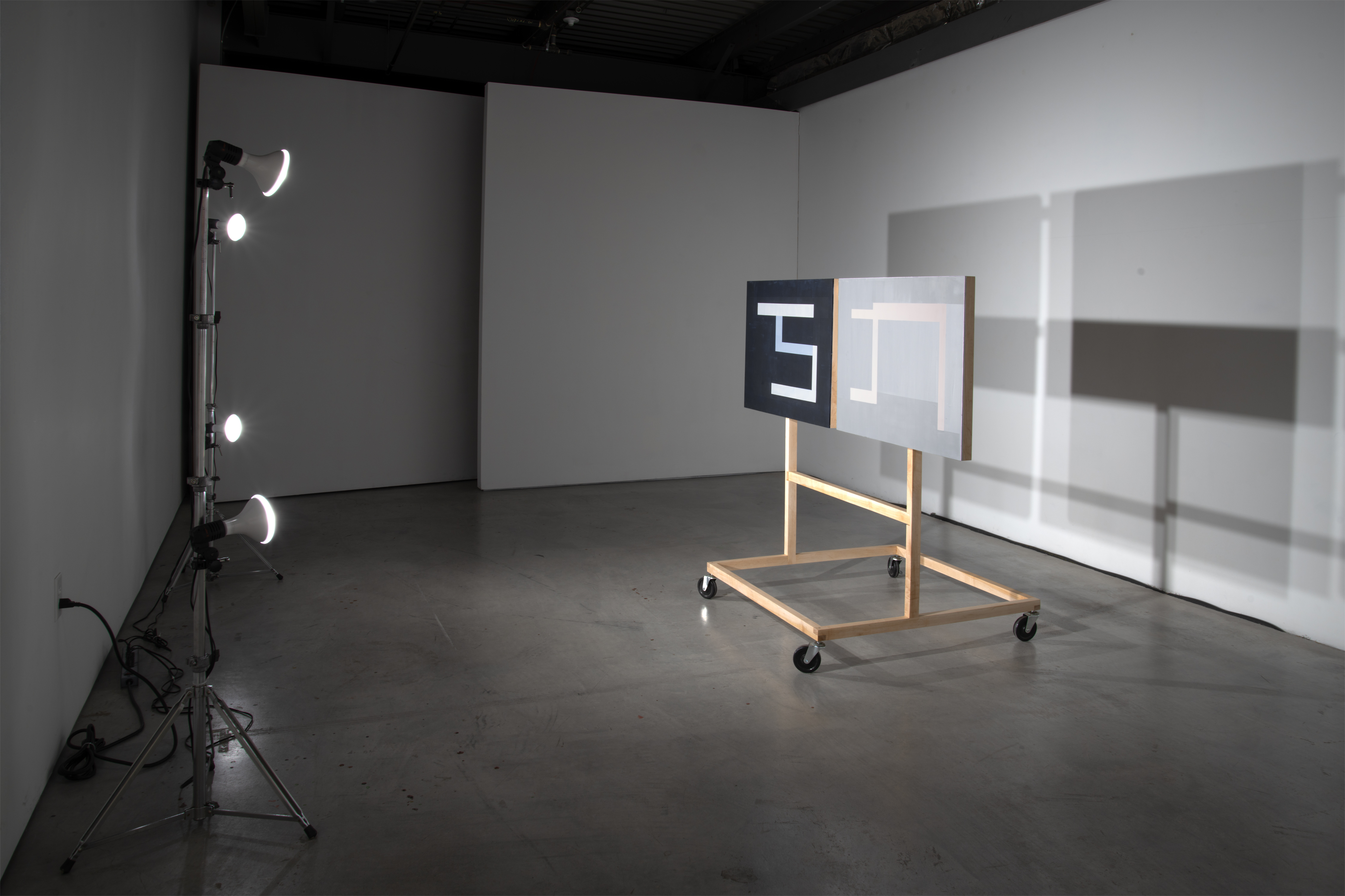 A wooden-framed mobile display on casters holds two abstract paintings—one predominantly black with white geometric forms, the other predominantly white with subtle architectural elements—positioned in an industrial gallery space lit by professional studio lights on a tripod.