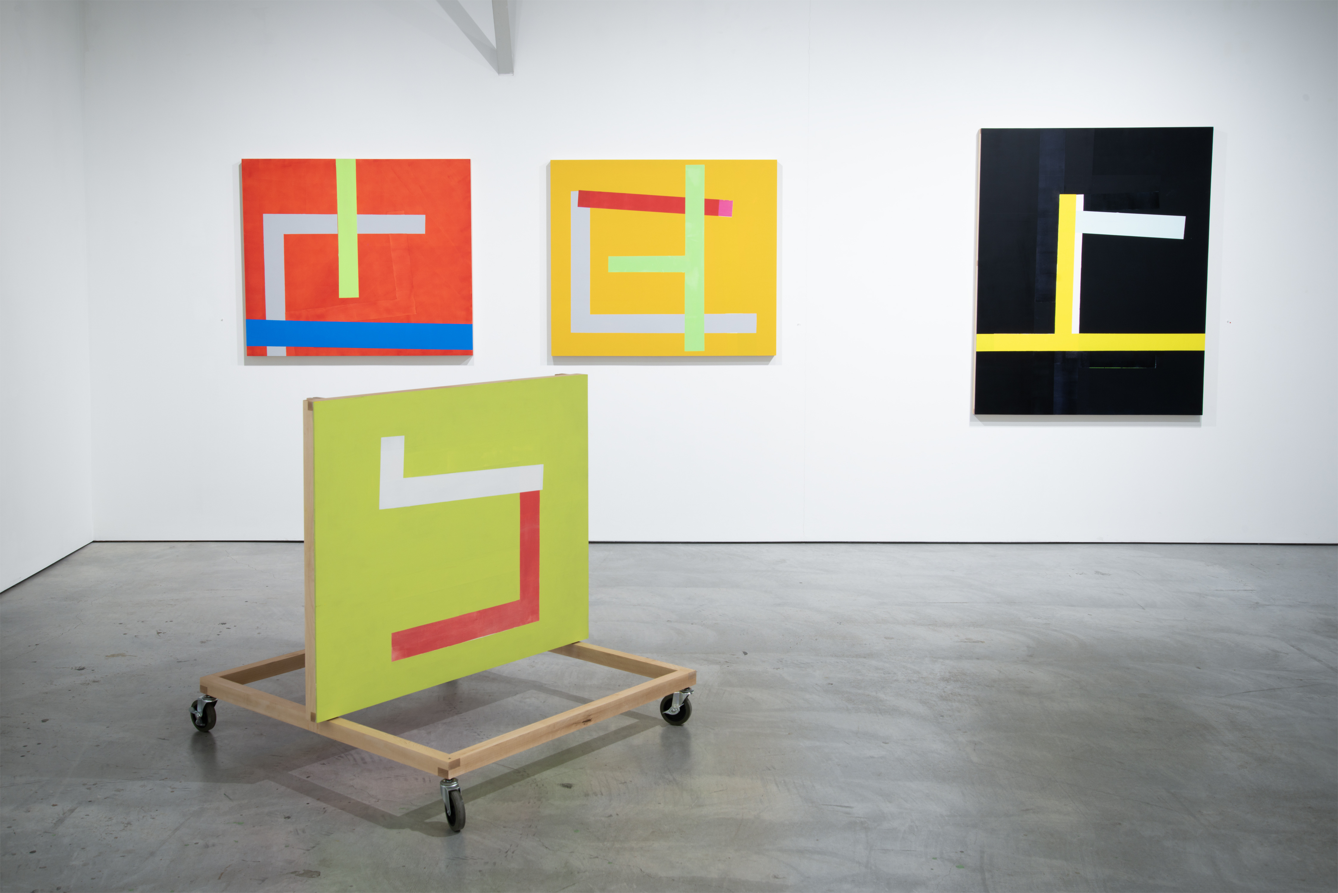 Four geometric abstract paintings displayed in a gallery space feature bold colored backgrounds—red, yellow, lime green, and black—with contrasting rectangular and linear forms in white, pastels, and bright accents. One lime green canvas is mounted on a wooden wheeled platform in the foreground, while three others hang on the white walls behind it.
